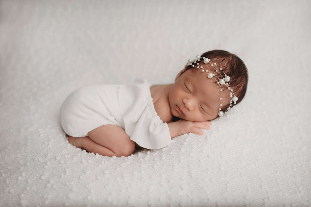 sleeping two week old baby girl wearing white off the shoulder romper with pearl headband lying on her tummy over textured white background