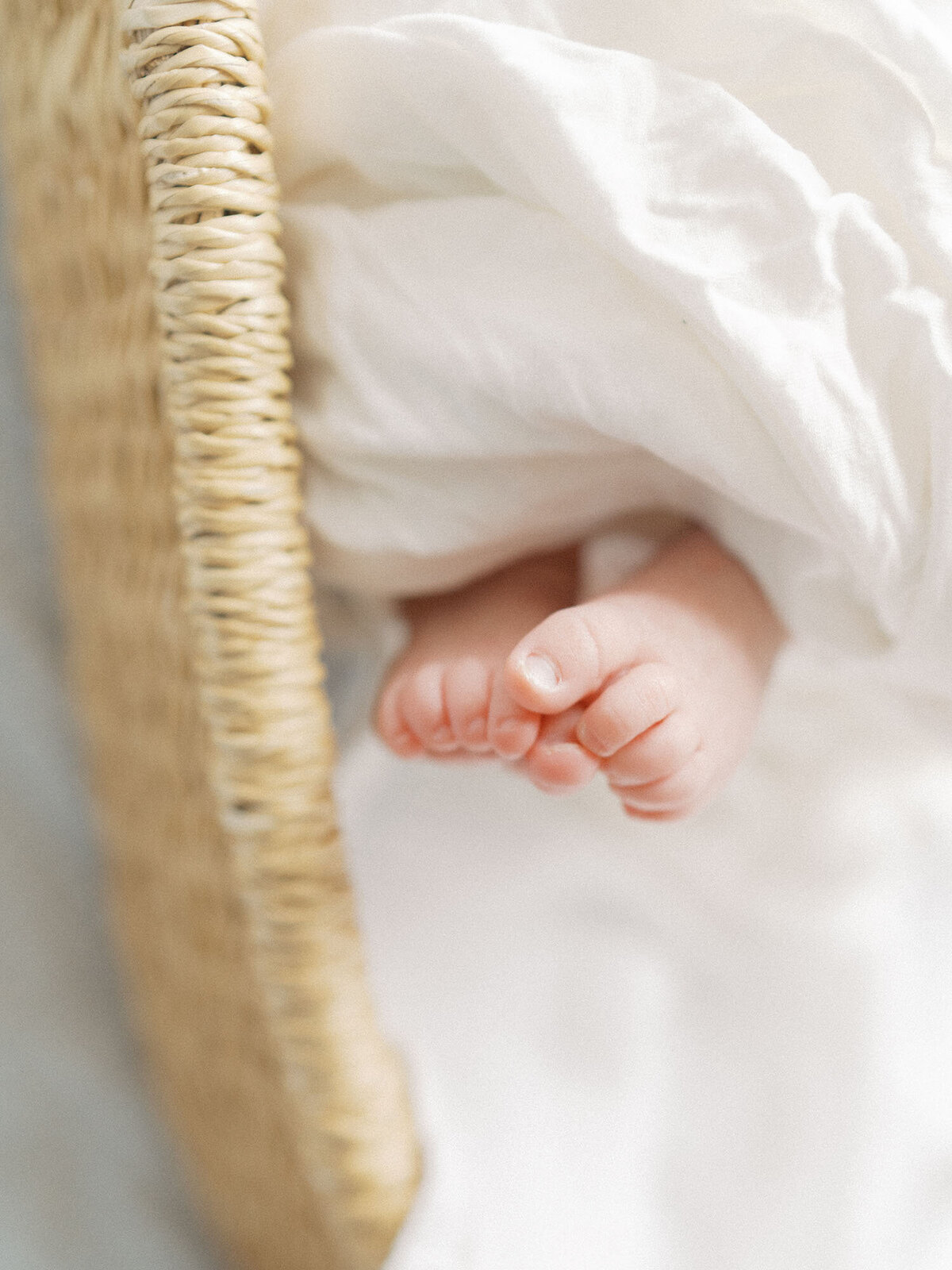A close-up of tiny newborn baby feet peeking out from a soft swaddle, resting gently in a woven basket. The delicate details capture the sweetness and fragility of newborn life