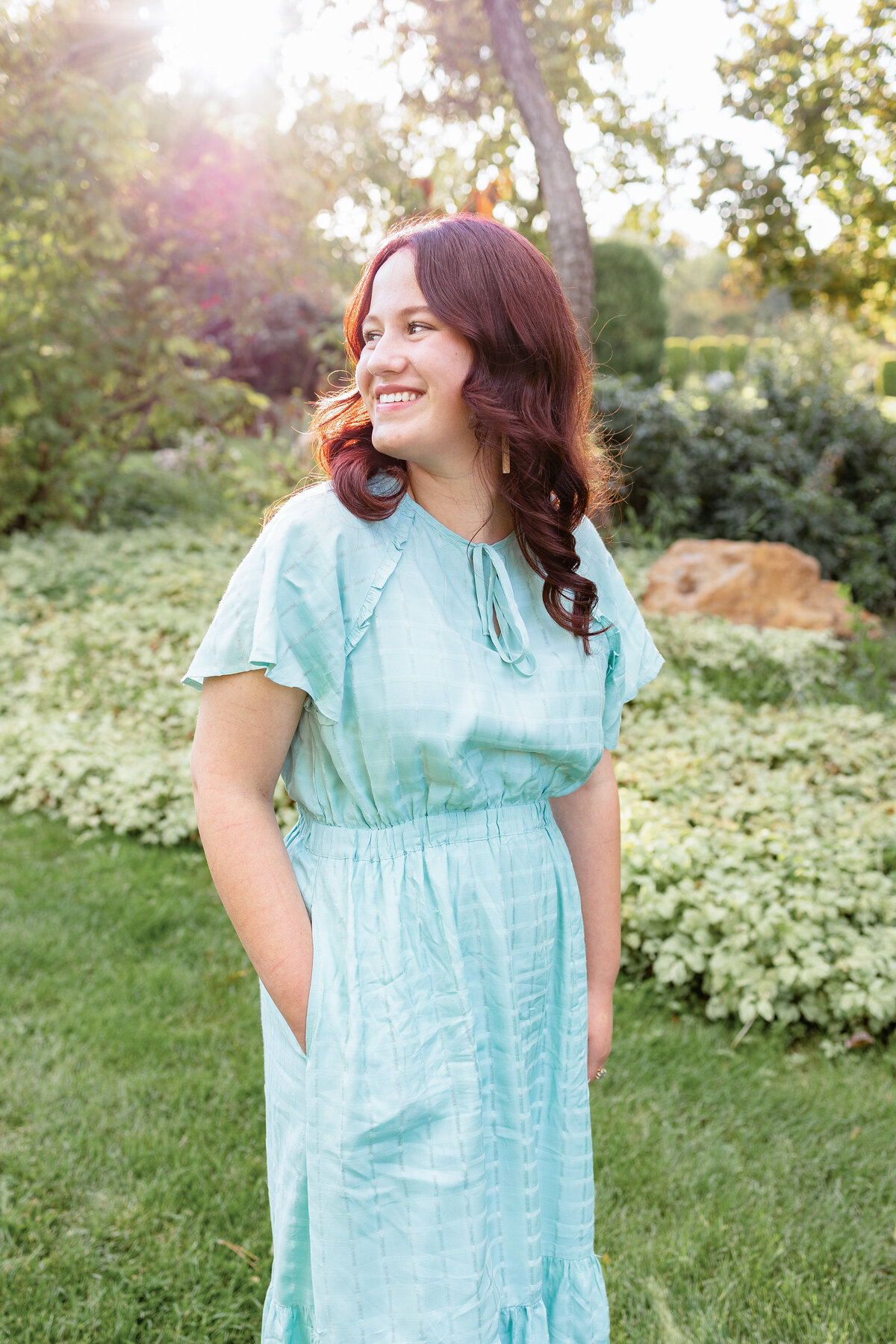 A young woman in a flowing blue dress smiles softly while standing in the golden afternoon light of a garden.