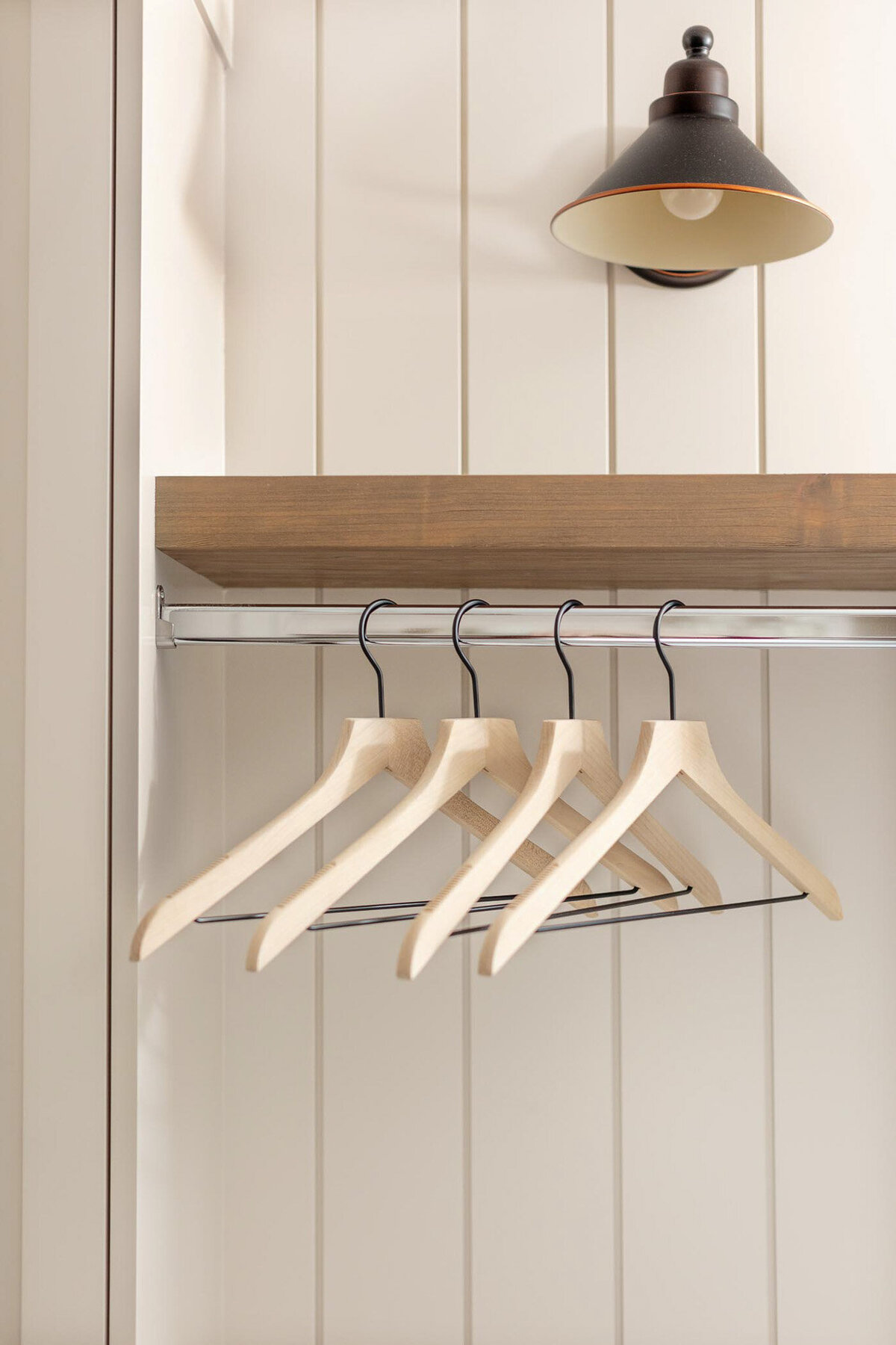 Shiplap wall painted creamy white with a natural wood floating shelf with a black sconce above. A silver rod under the floating shelf with wooden hangers on it.