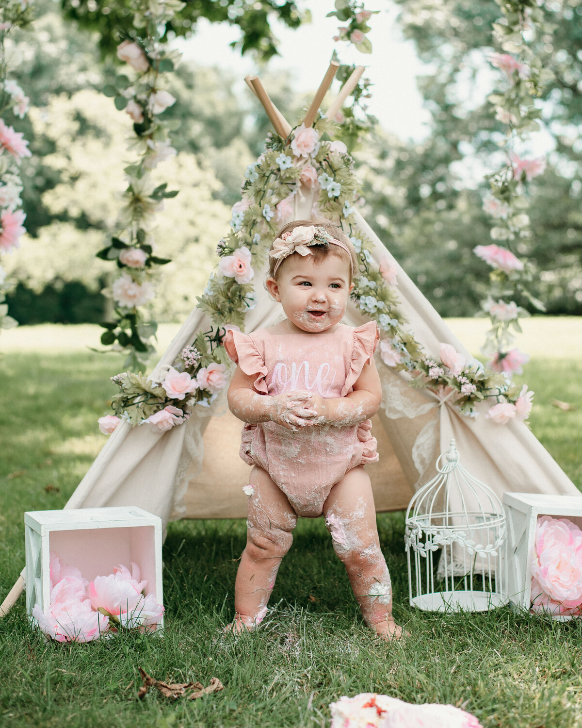 One year girl in pink ruffle sleeve romper standing in grass in front of floral tee pee with icing on her hands, feet and legs