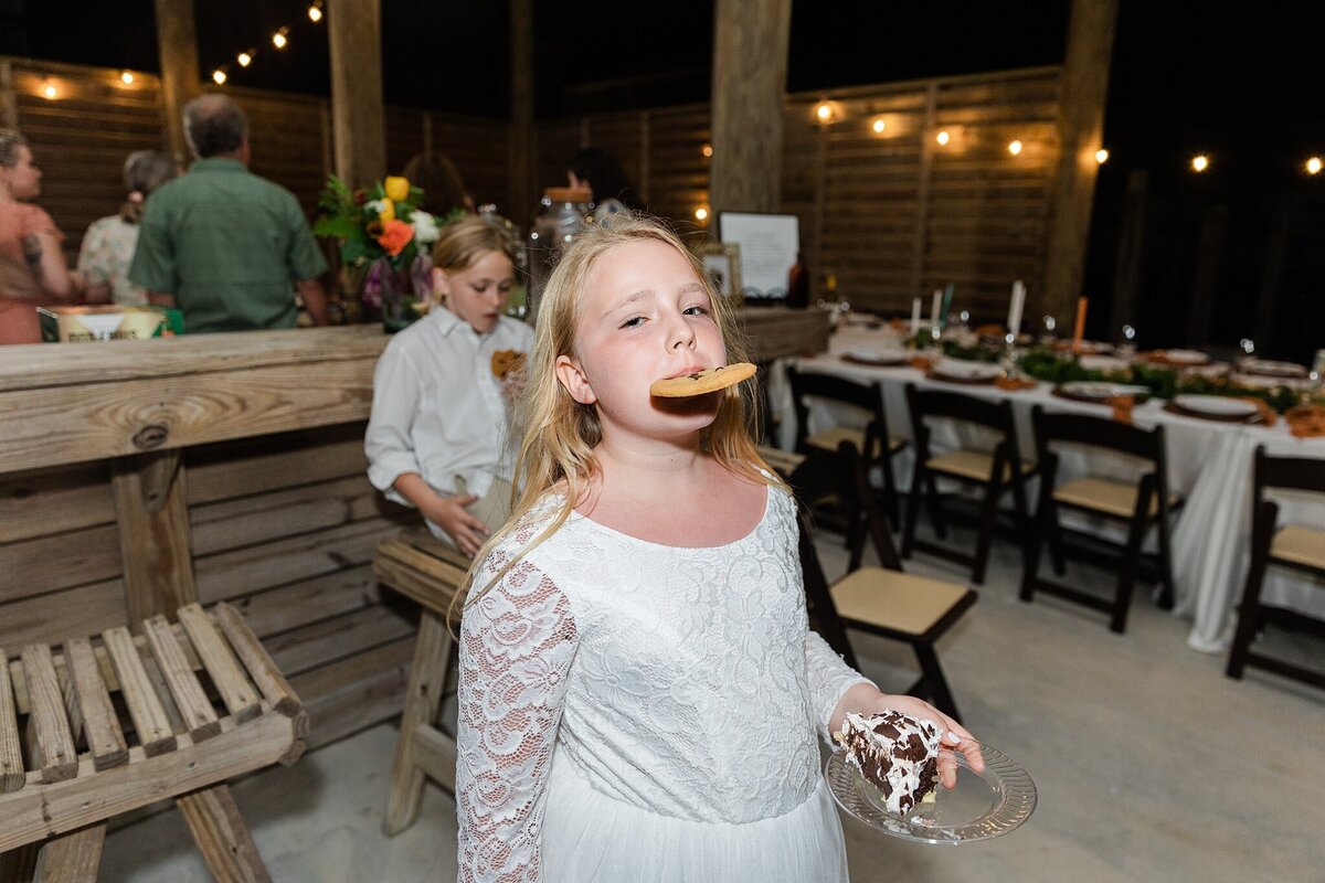 A playful photo of the groom's daughter enjoying the dessert offerings at a wedding reception in Crystal Beach, Texas. The daughter is wearing a long sleeve, intricate, white dress and holds a piece of cake in one hand while holding a cookie directly in her mouth.