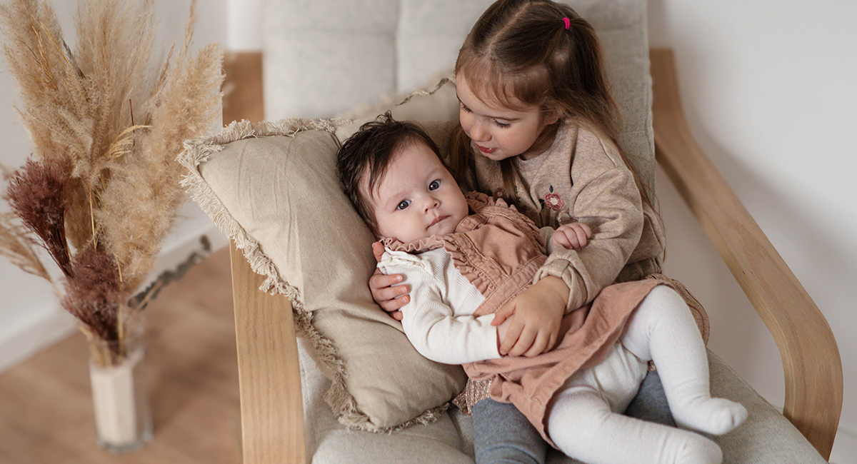 Die große Schwester sitzt auf einem beigefarbenen Sessel und hält ihre kleine Schwester in den Armen
