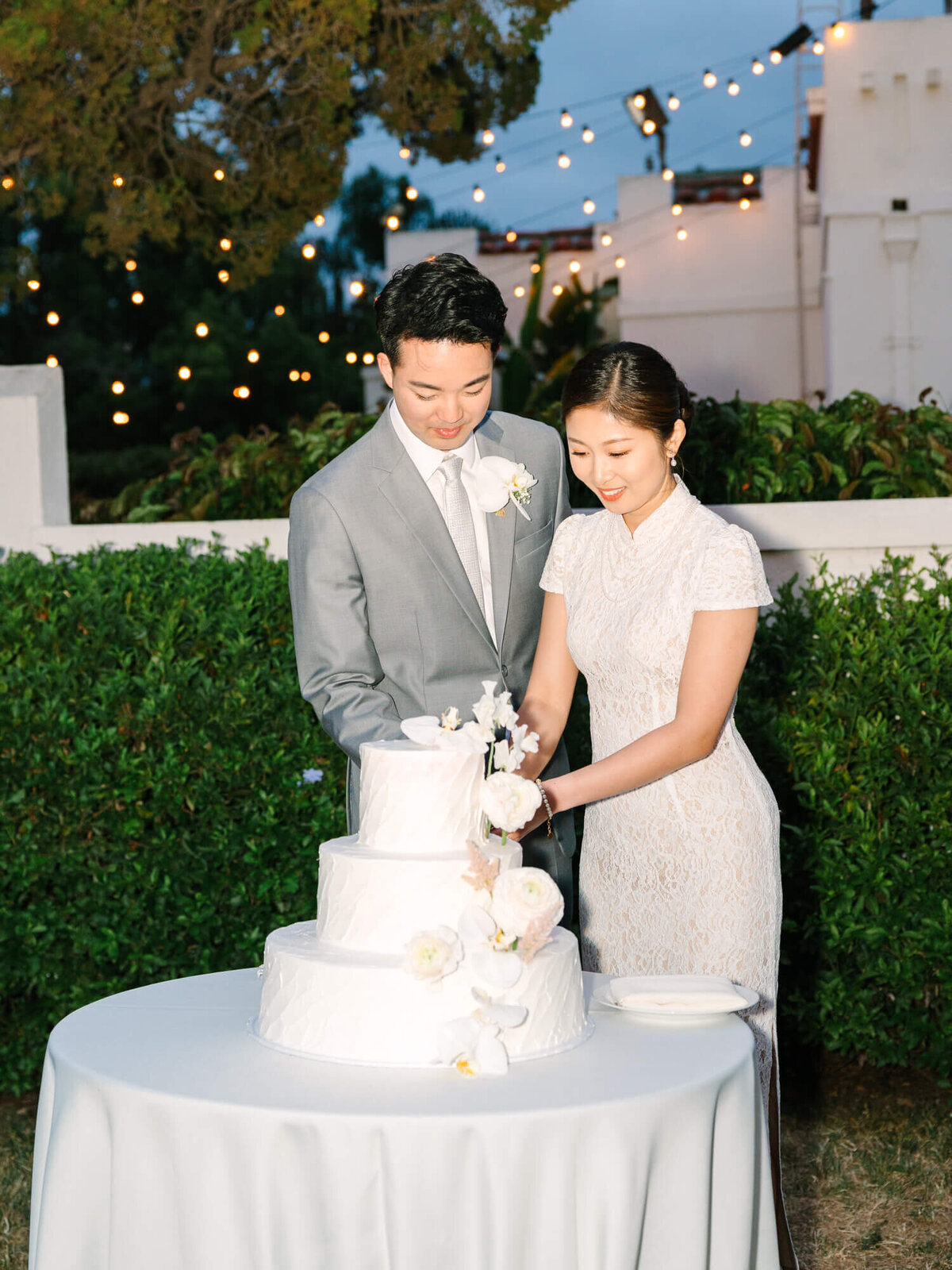 A couple in suit and Chinese wedding gown cuts a white, tiered cake adorned with flowers. They are outdoors, with elegant string lights and greenery in the background.