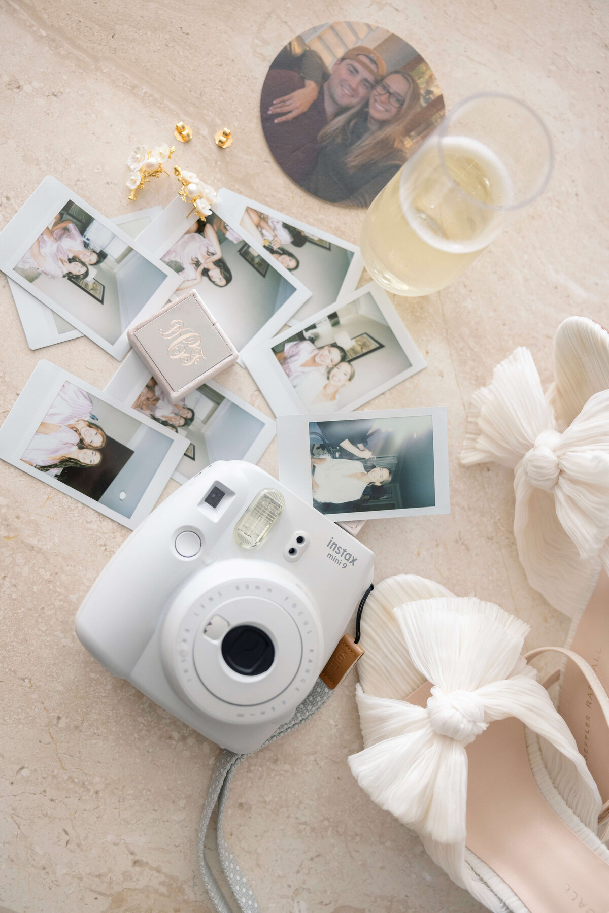 A white instant camera surrounded by polaroid photos, a pair of white heels with bows, a round photo of a couple, jewelry, and a glass of champagne on a light marble surface.