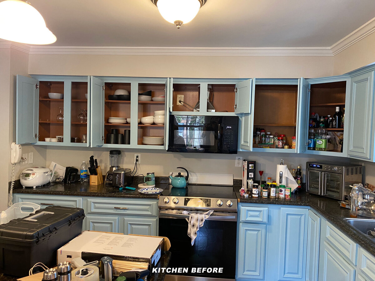 A kitchen with open blue cabinets displaying dishes and pantry items. A stove with a kettle and a microwave are visible. Countertops hold various appliances and boxes. A ceiling light illuminates the space. Text reads "KITCHEN BEFORE.