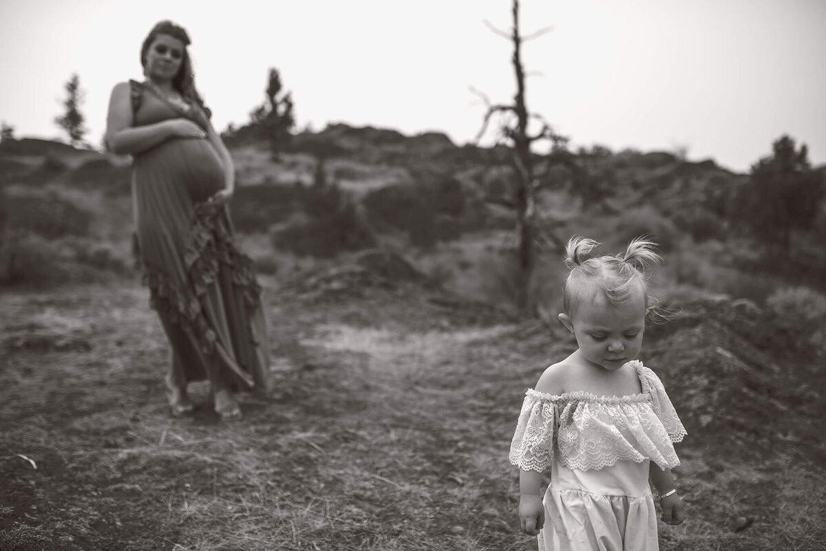 A black and white motherhood portrait. A pregnant mother stands in the background looking at her toddler daughter in the foreground who is wearing a long white dress and looking down at the ground. Taken by Brittany Danielle in Kamloops.