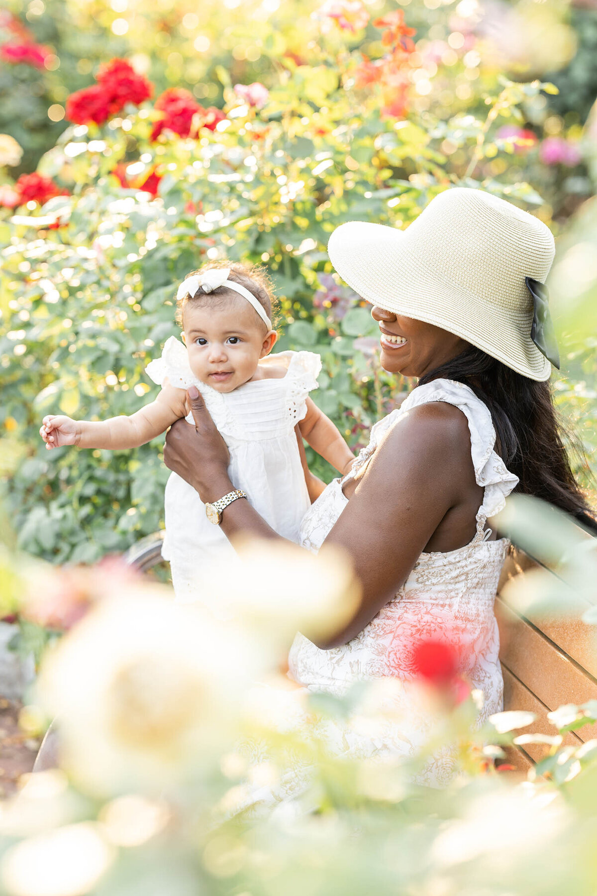 Baby girl in white dress is standing on mom's lap as mom sits on a bench in a rose garden during a mommy and me session in Connecticut