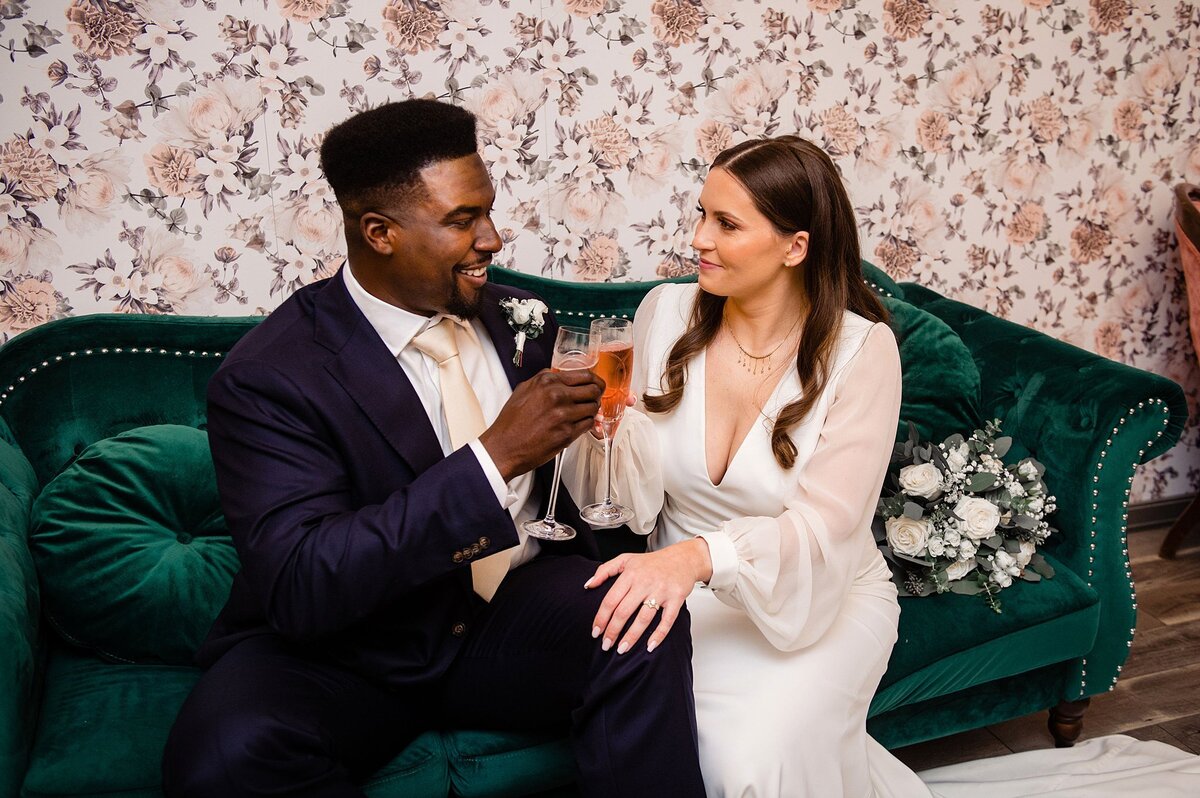 African American groom wearing a dark blue suit clinks champagne glasses with the bride wearing a long sleeve wedding dress with a plunging neckline. The bride and groom are sitting on an emerald green couch in front of a floral wallpaper wall. The white and green bridal bouquet sits on the green sofa behind her at Steel Magnolia Barn.