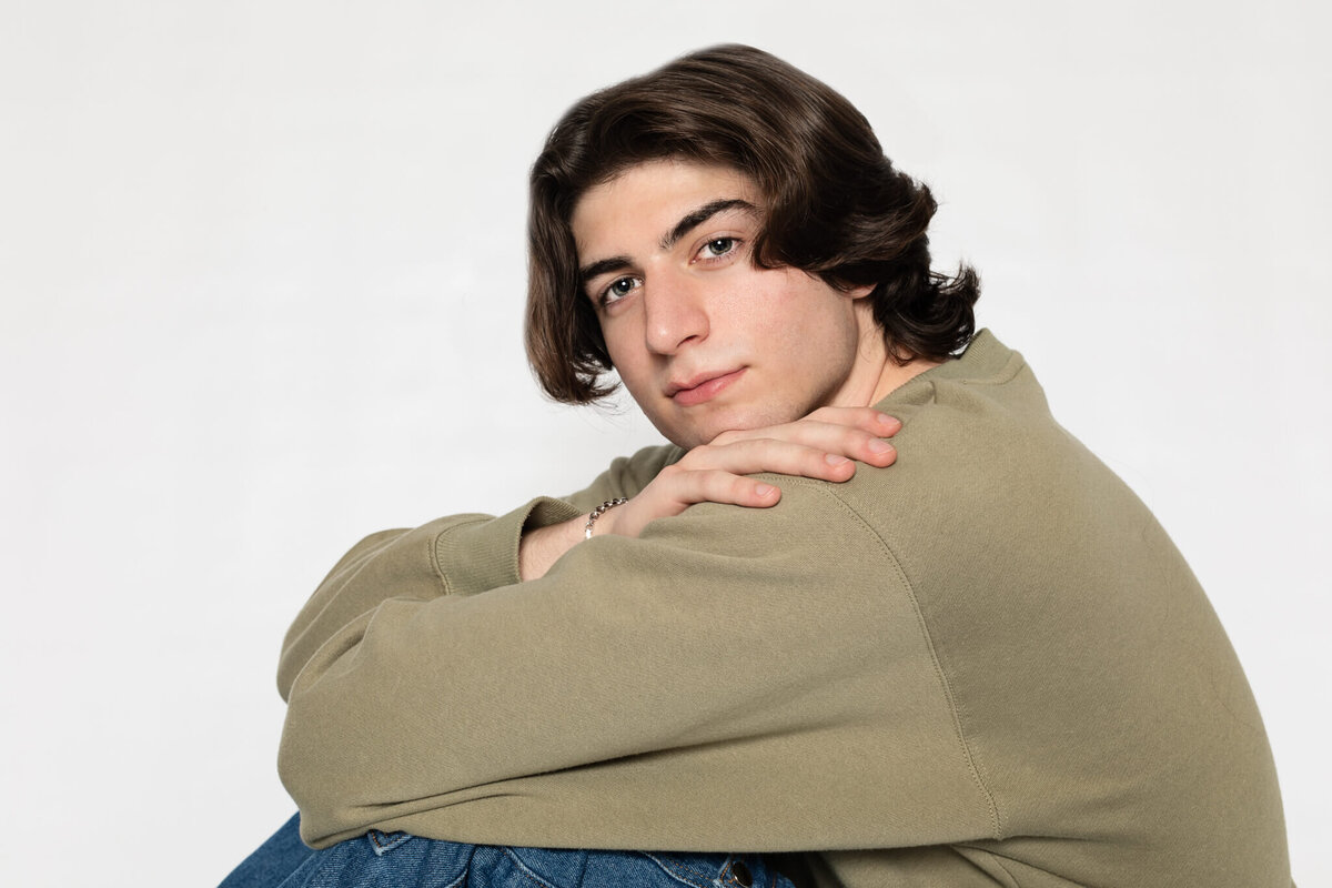A person with shoulder-length dark hair wearing a green sweater and jeans sits with arms crossed over knees against a plain white background, looking at the camera.