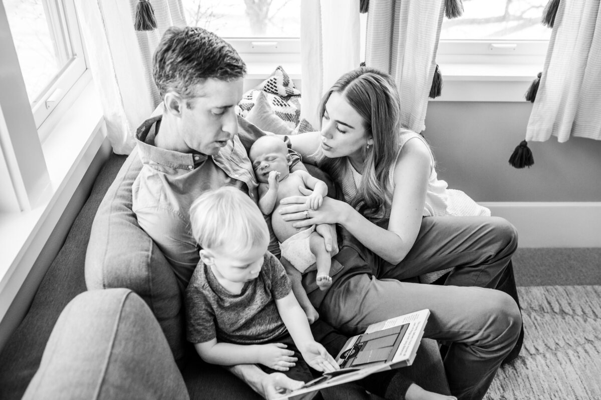 A family of four sits on a couch; the father reads a book with the toddler while the mother lovingly holds the newborn.