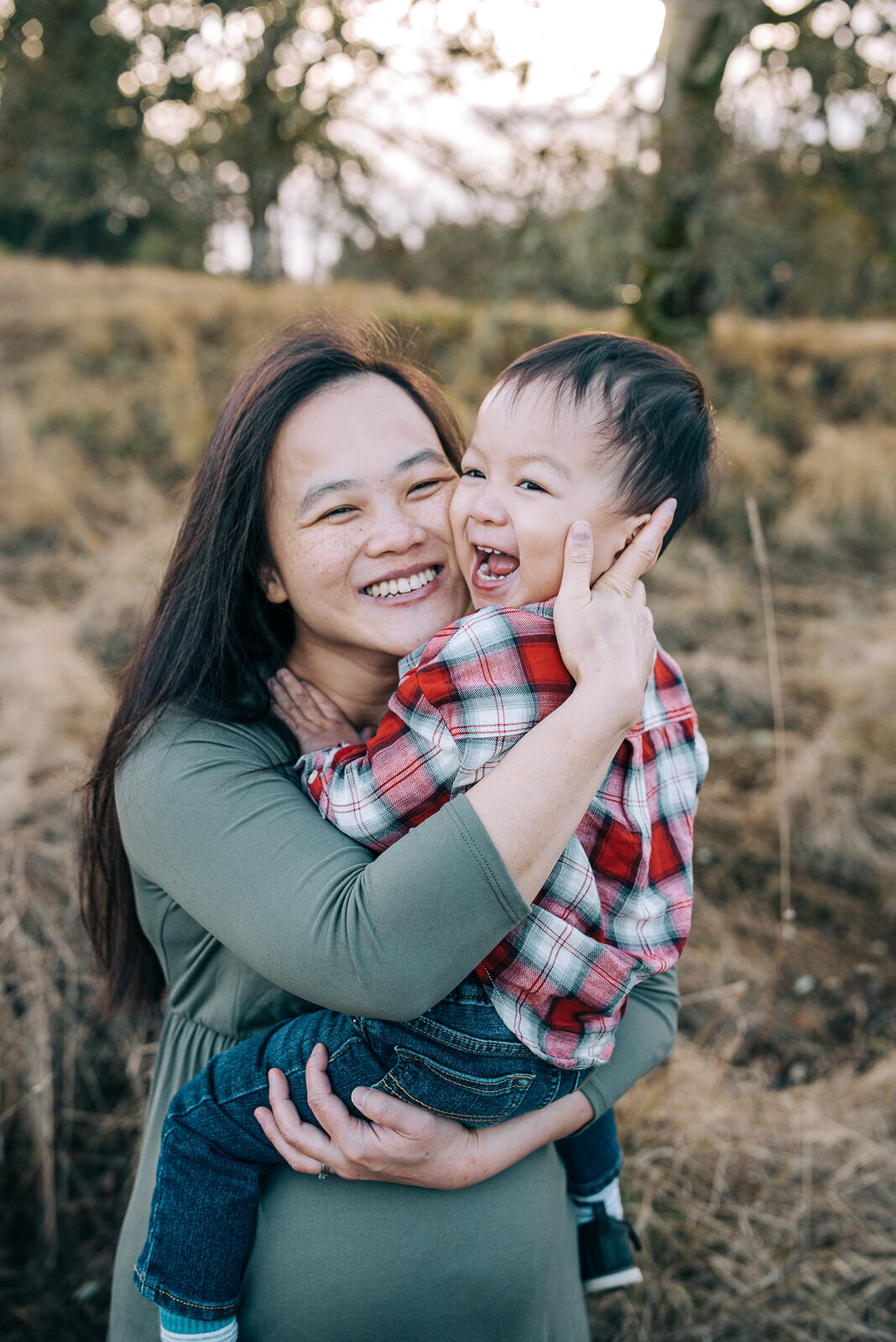 Mom hugging her toddler son while he sits on top of her pregnant belly in field at Fort Steilacoom park in Lakewood