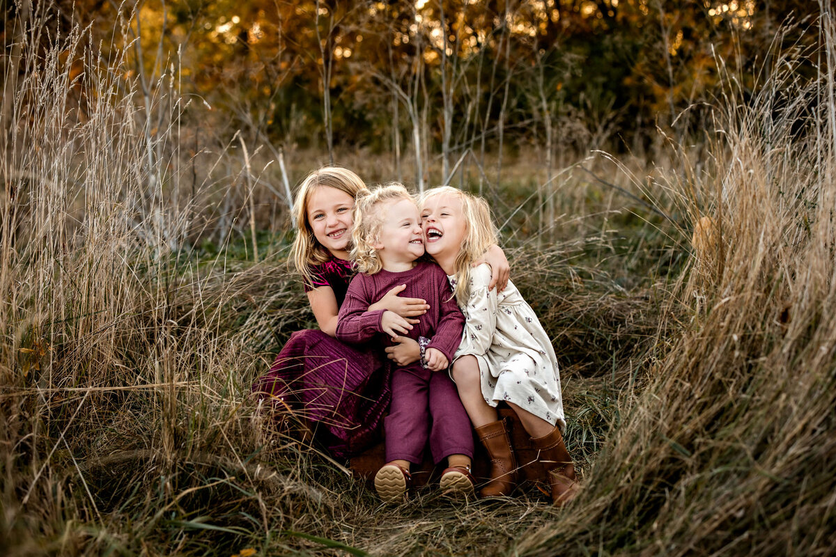 Three young children sit together in a grassy field, laughing and hugging. They are surrounded by tall grasses with trees in the background, creating a warm, autumnal setting.