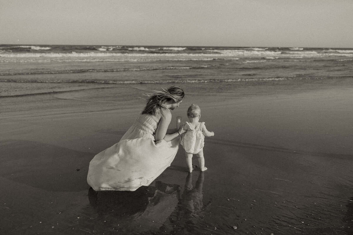 Black and white photo of a mother and toddler daughter walking hand in hand along the shore during a Charleston beach family session.