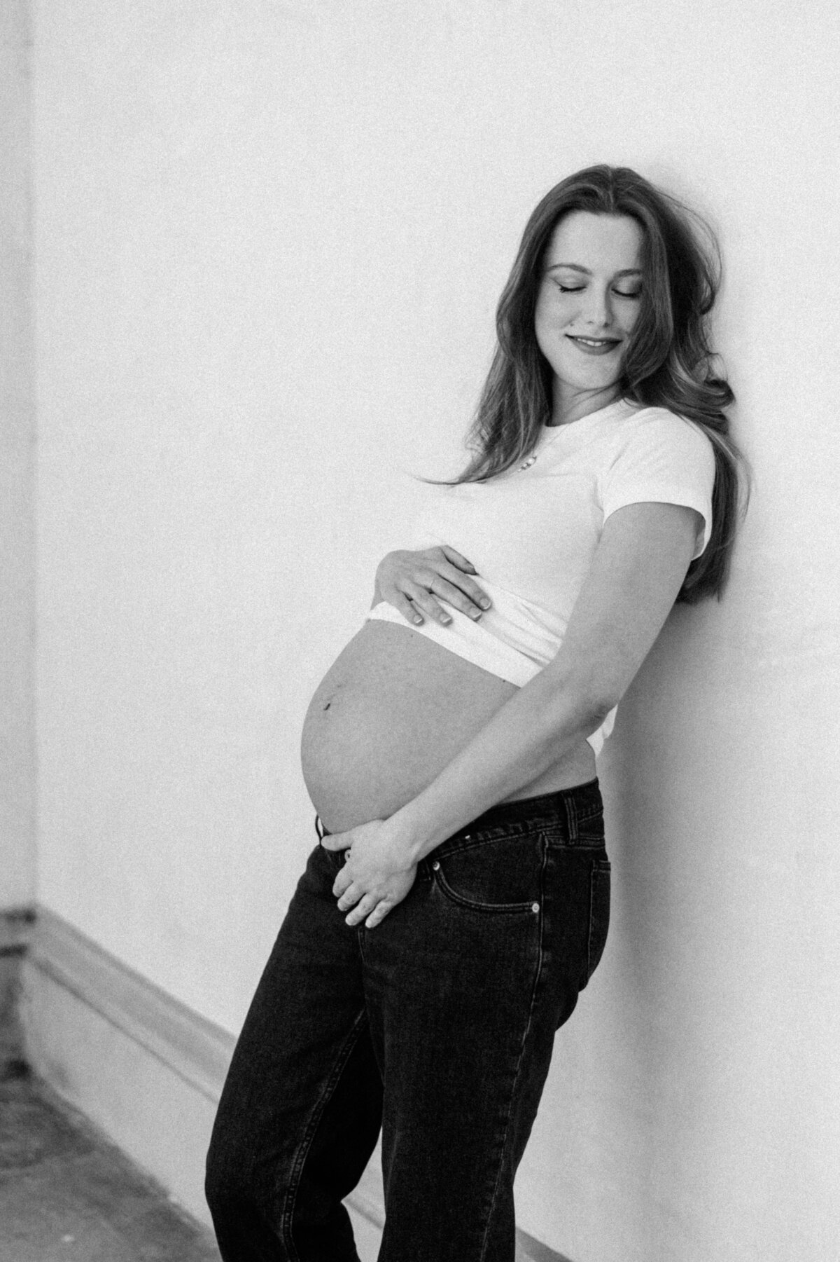 Black-and-white maternity portrait of a smiling first-time mom leaning against a studio wall, wearing a cropped tee and jeans