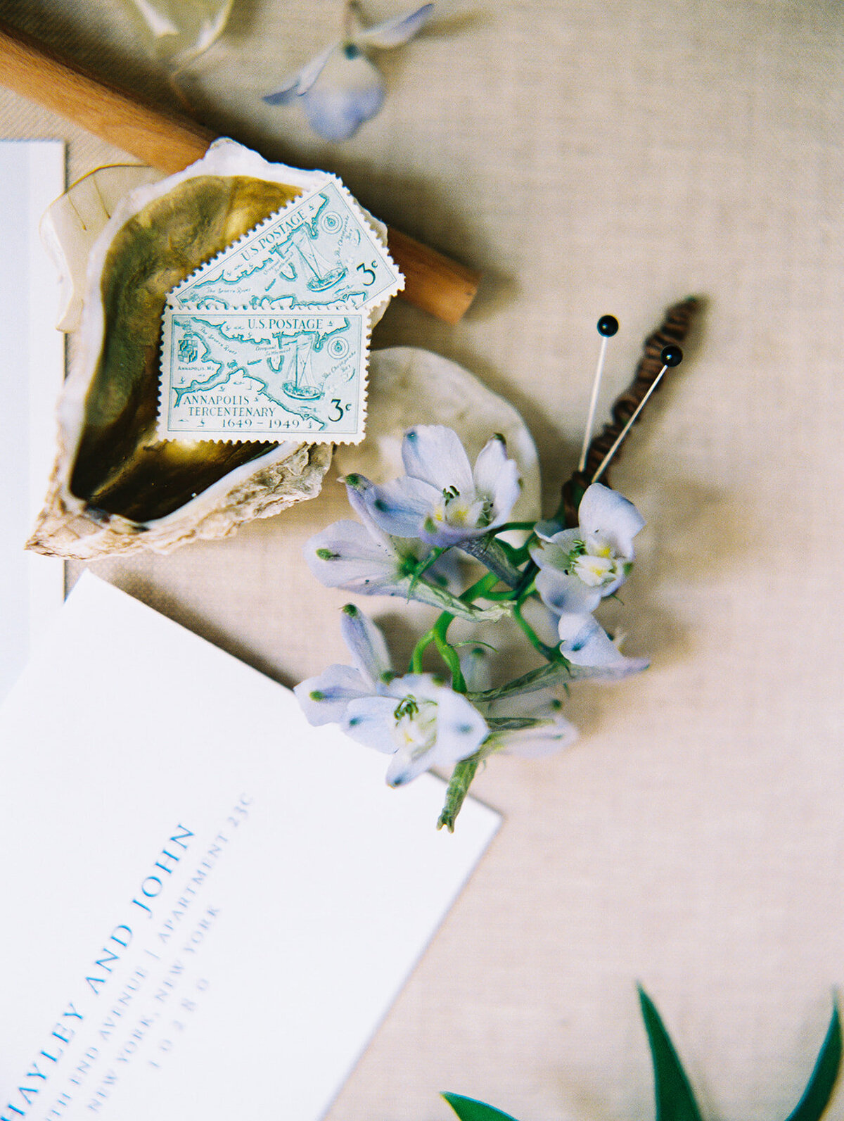 The photo was taken at The Hamptons, New York, waterfront estate tented wedding by photographer Bonnie Sen. It features a flat lay image showcasing two vintage postage stamps, blue flowers, a seashell, a pencil, and part of a wedding invitation with HAYLEY AND JOHN visible. This charming arrangement is elegantly set against a neutral fabric background.