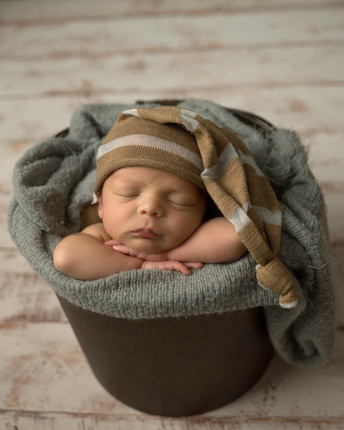 Newborn boy with brown and white sleepy hat sitting in a pail sleeping.