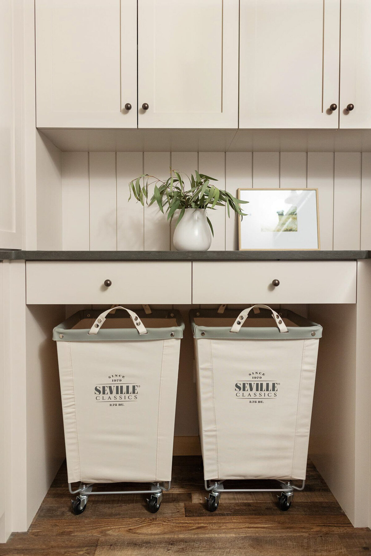 Laundry room with dark brown wood floors. Open shelving area with upholstered laundry hampers on casters. Beige cabinetry and shiplap walls. Beige cabinet with doors with antique bronze knobs. Grey quartz countertops above with open shelving painted beige. Shelves adorned with glass jars of laundry detergent and pods, cleaning spray bottles, towels, jars, and wicker baskets.