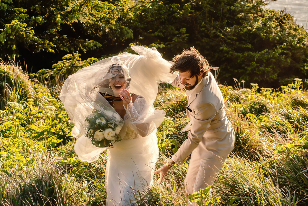 during their Oregon coast elopement, a bride and groom in their wedding attire, explore a grassy, coastal cliff. The smile wide as the wind whips the brides dress and her groom attempts to ccatch the layers of her dress.