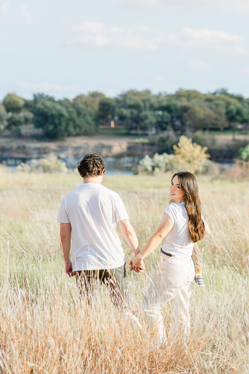 A mother and father walk through a field of golden grass at an Austin park while the mother looks back at the photographer.