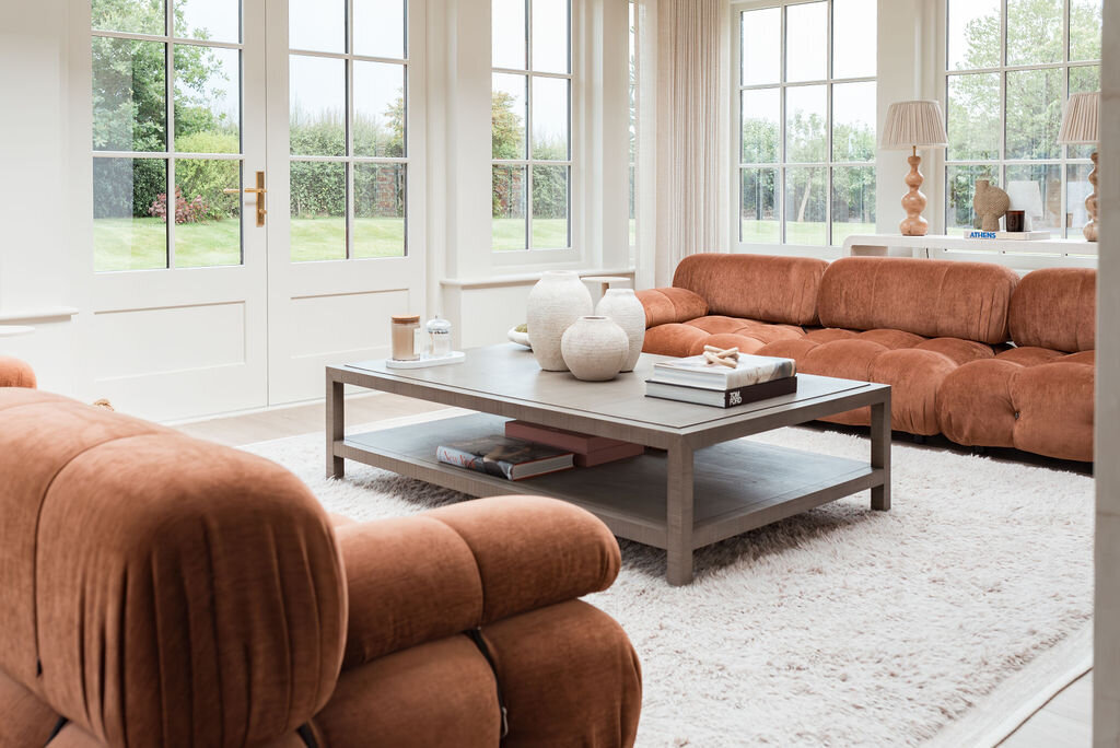 A cozy living room with large windows showcasing a lush green garden outside. The room features two rust-colored, plush sofas, a light grey coffee table with vases and books, and a soft, white rug covering the floor. Two lamps are on side tables flanking a large sofa.