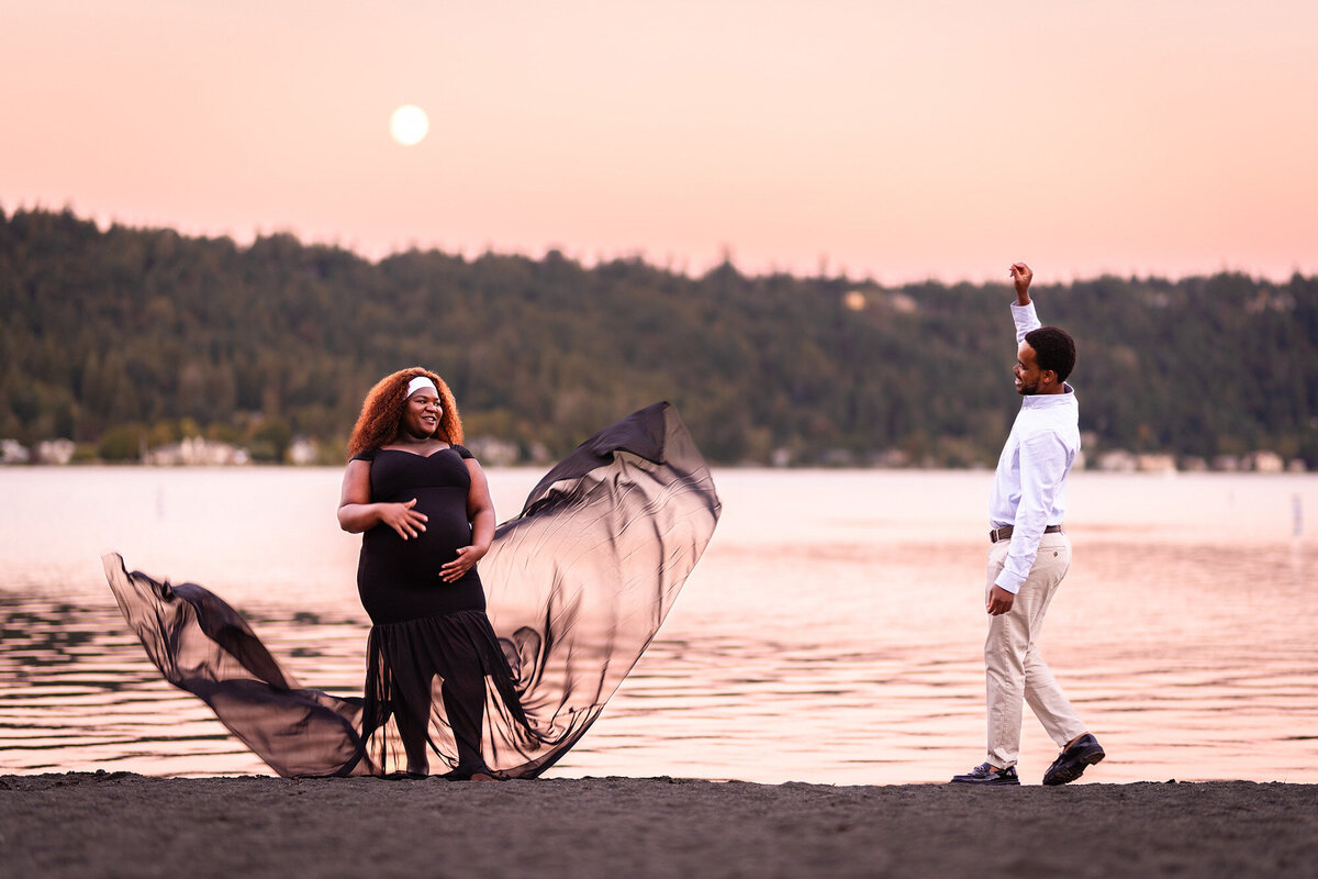Maternity photoshoot taken from golden light into twilight hours in Seattle, Washington. The photograph shows a dramatic portrait of an expectant woman and her husband tossing the train of her flowing maternity gown into the air. The image freezes the dress in mid-air, framing her baby bump and creating a cinematic wind-blown effect against the Seattle night sky.