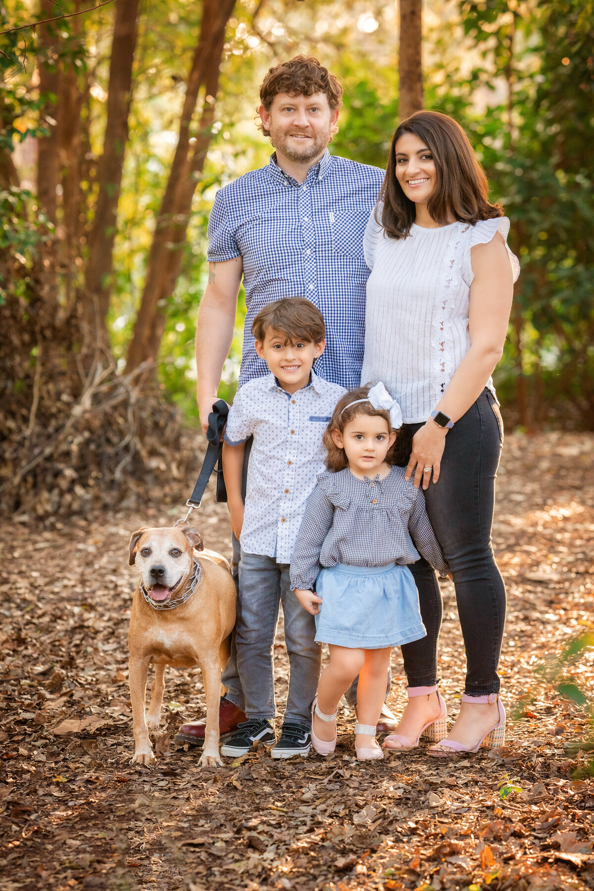 Mom, dad, son, daughter, and dog are standing in the park.  There are brown leaves on the ground and trees behind them.  Mom is wearing a white blouse and black jeans.  Dad is wearing blue.  The kids are wearing jeans and a skirt.  The dog is tan colored and on a leash.  They are all looking at the camera.