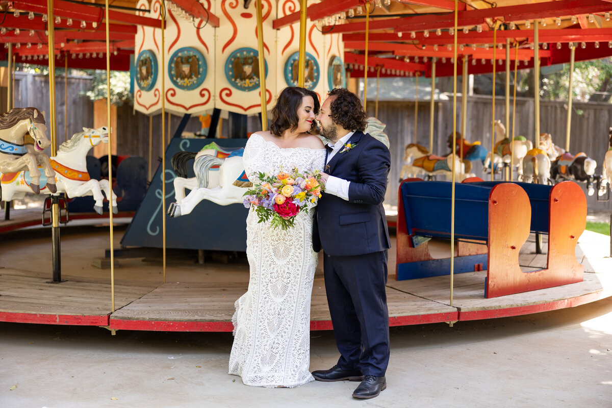 A newlywed couple about to kiss in front of a merry go round