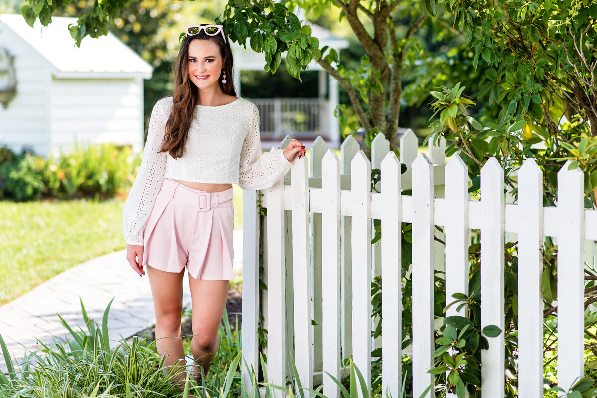 Monacan high school senior girl wearing pink shorts and white shirt poses against picket fence at Amber Grove for her senior portraits.