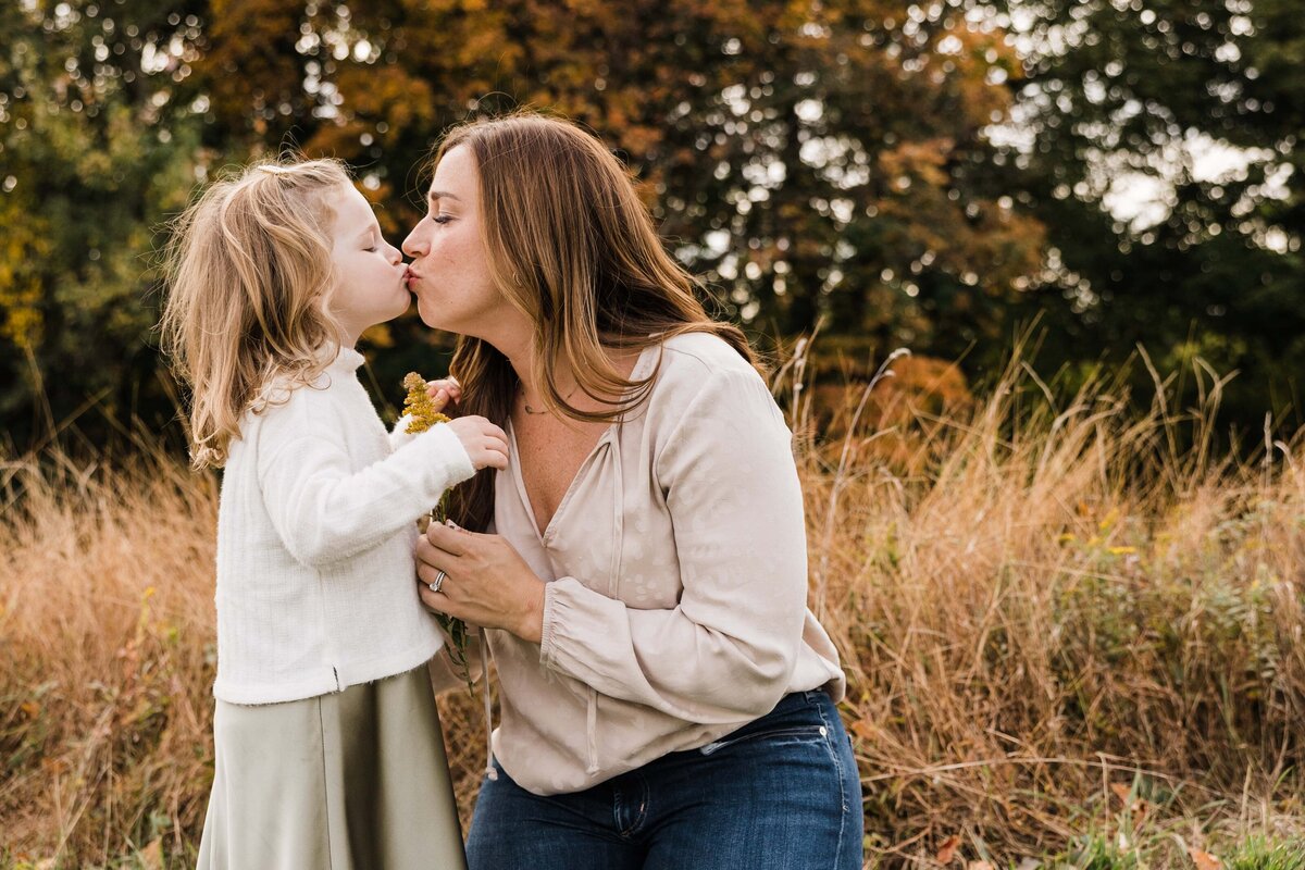 A young girl and a woman sharing a tender moment with a kiss on the lips in an outdoor setting with autumnal foliage, captured beautifully by a family photographer in Pittsburgh.
