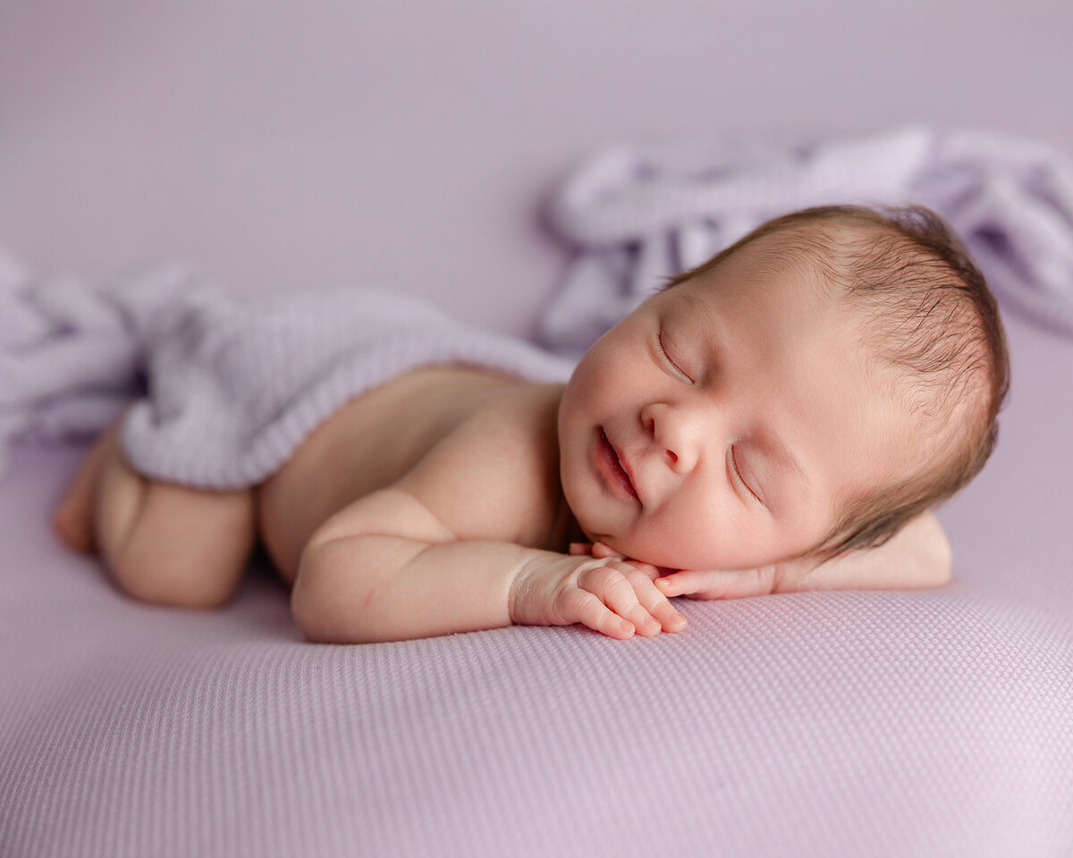 Newborn baby boy smiling in his sleep, comfortably posed on a soft lavender backdrop with a gentle swaddle. Captured during her Inland Empire session by Kaitlyn Dawn Photography.