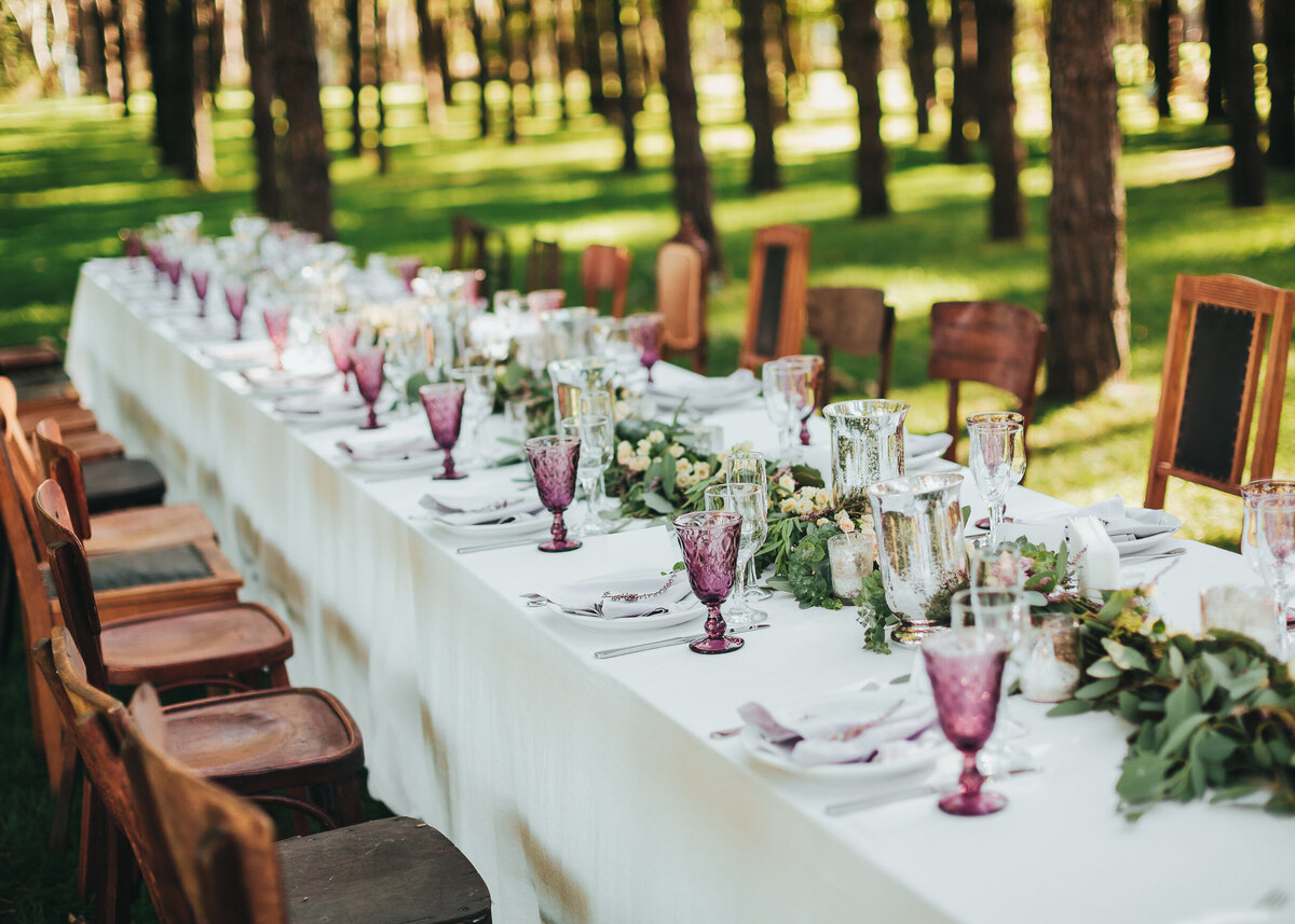 Event table decorated with branches of greenery, stands on green grass in a wood, ready for the wedding party.