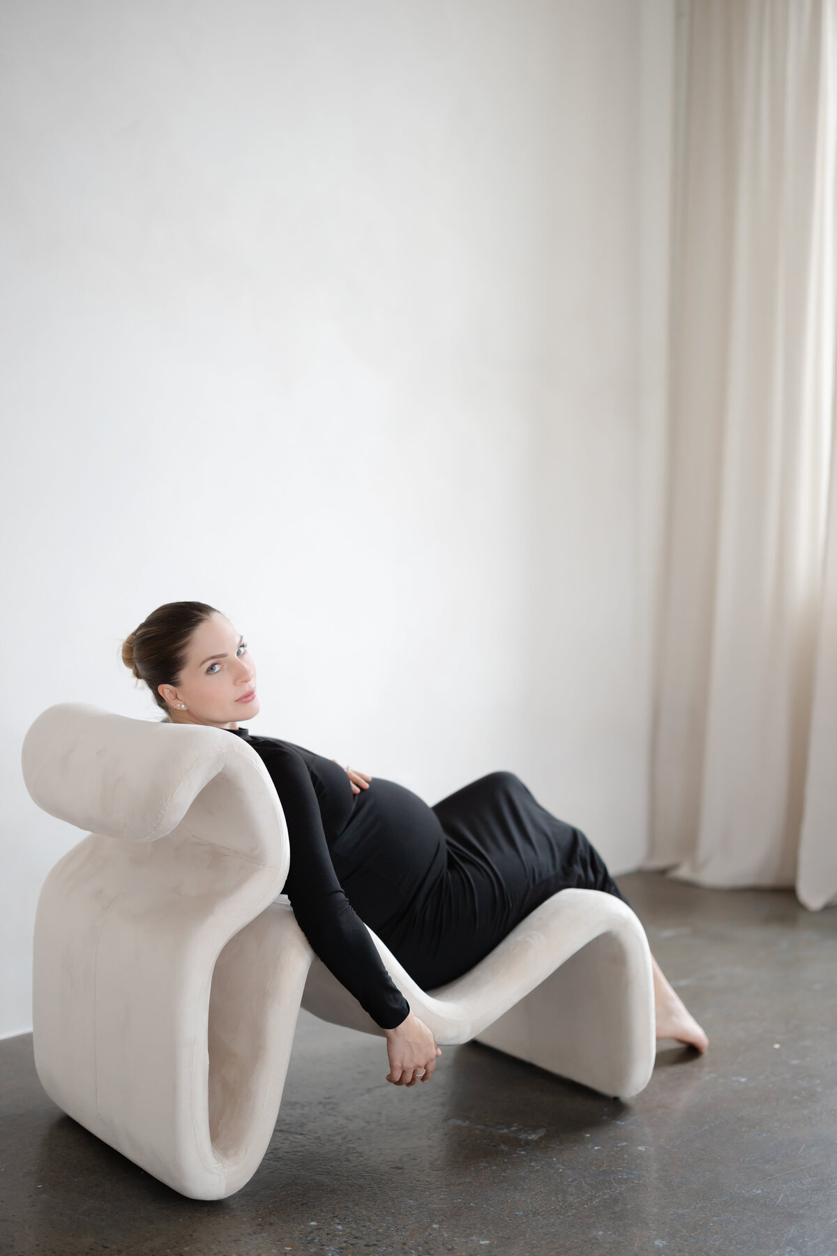 a woman wearing a black dress sits in a funky and modern beige chair as she holds her pregnant belly and glances back at the camera