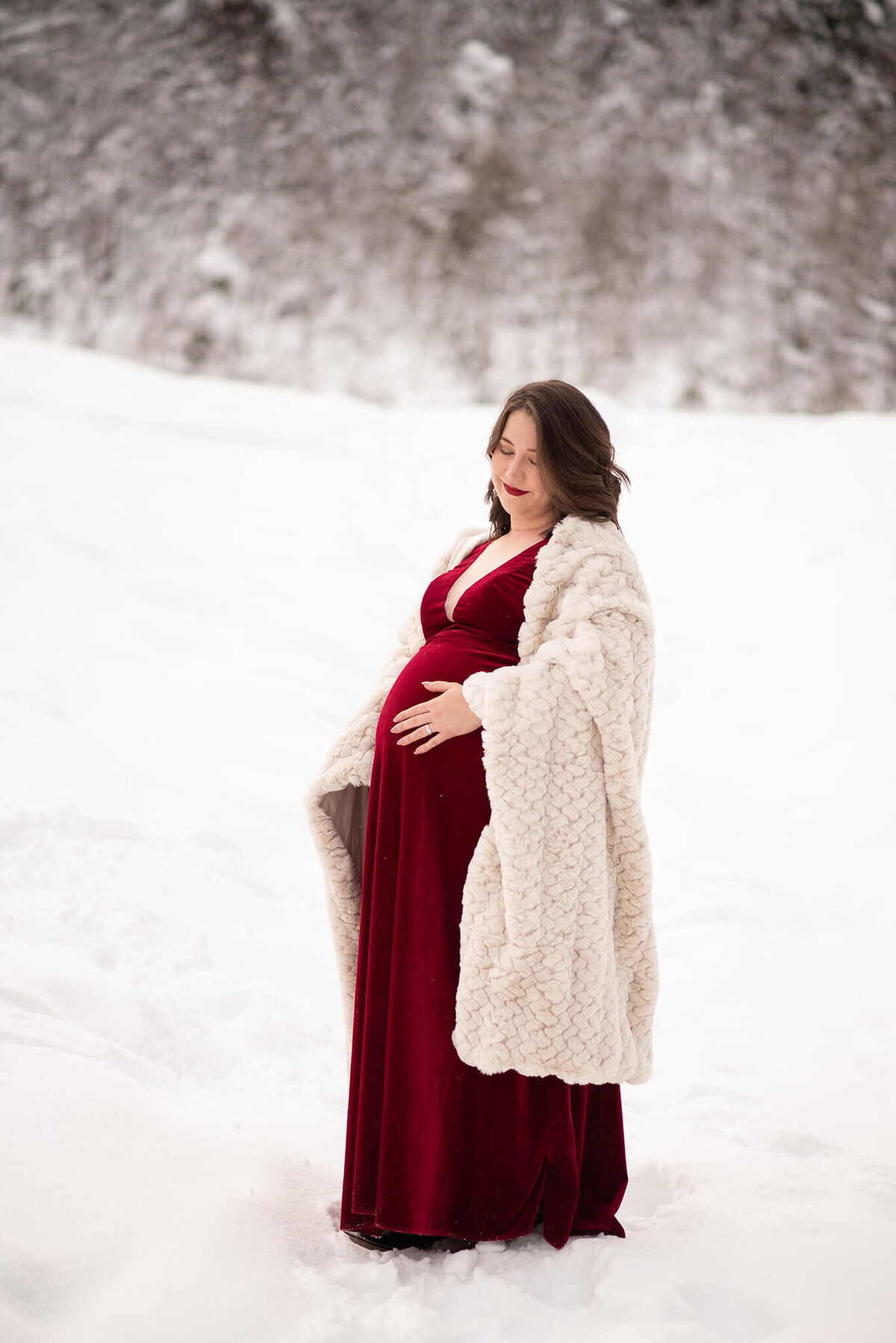 A woman stands in the snow under a fuzzy blanket. She is pregnant and wearing a red velvet dress. She holds her bells. Taken by Brittany Danielle