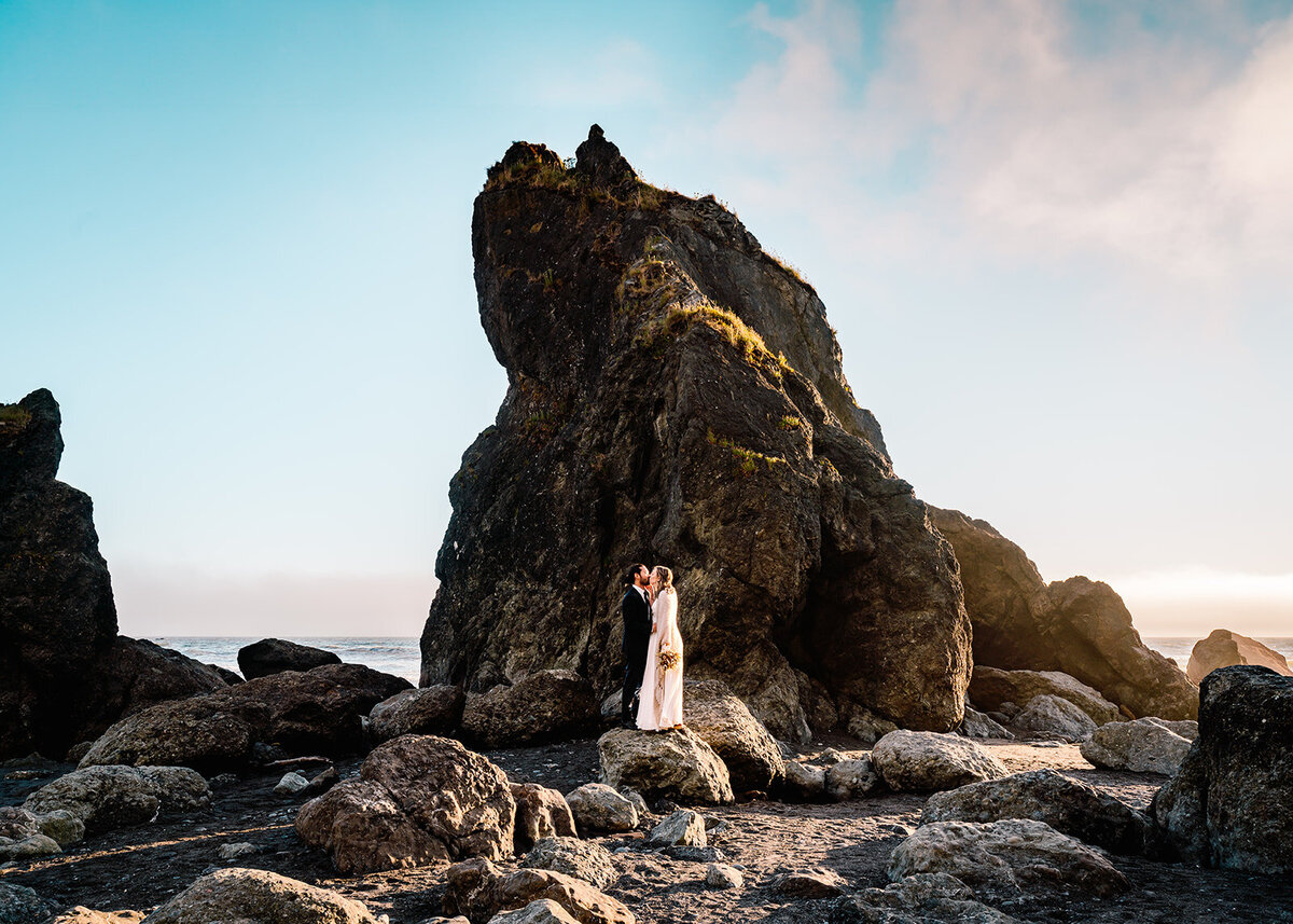 during a sunset elopement package, a bride and groom in their wedding attire kiss in front of a seastack. The rocks are highlighted by the setting sun and we see a bright, blue sky above them with soft gold clouds rolling in