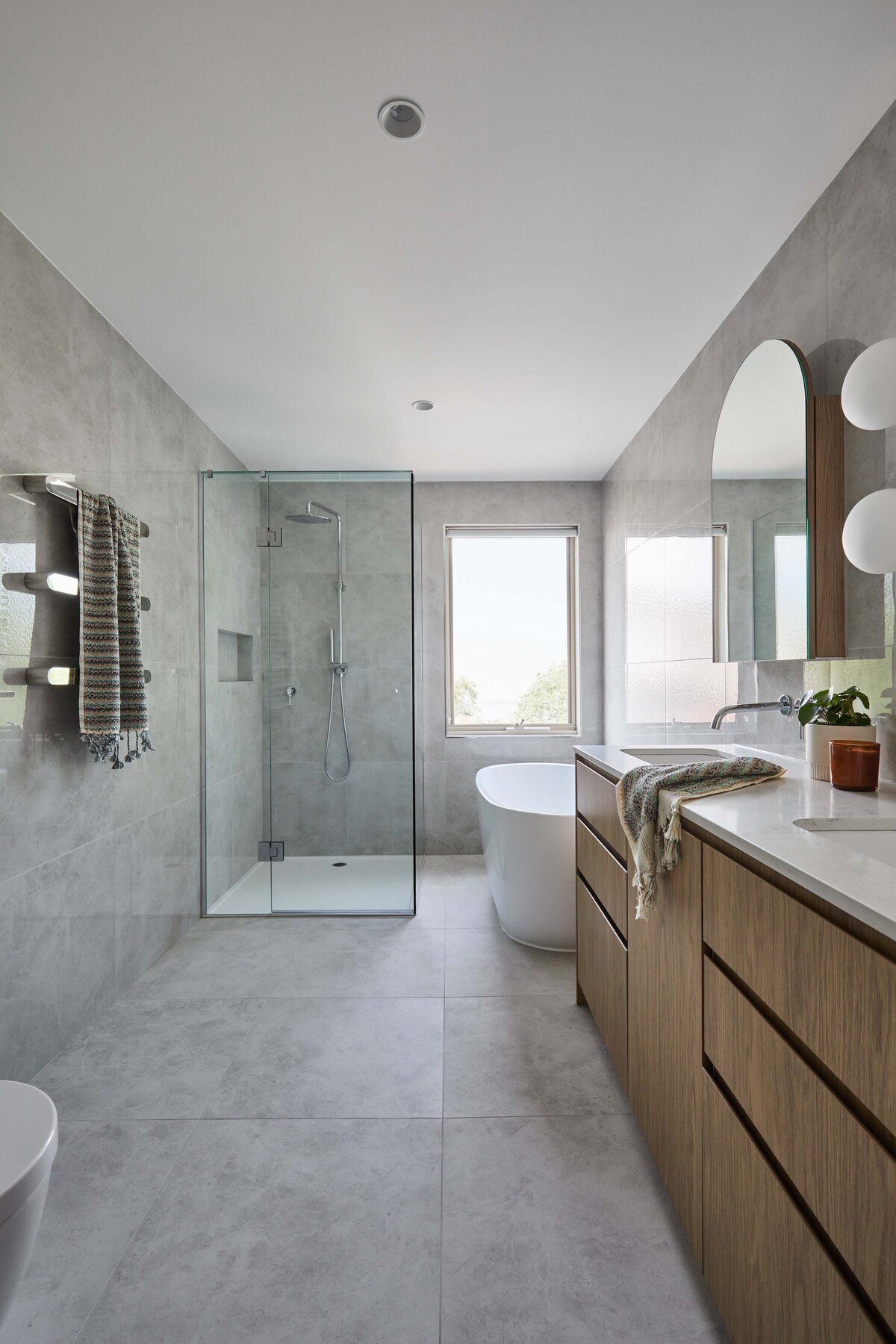 Modern bathroom with large format grey tiles, timber drawers, freestanding white bath and glass shower with chrome fittings.