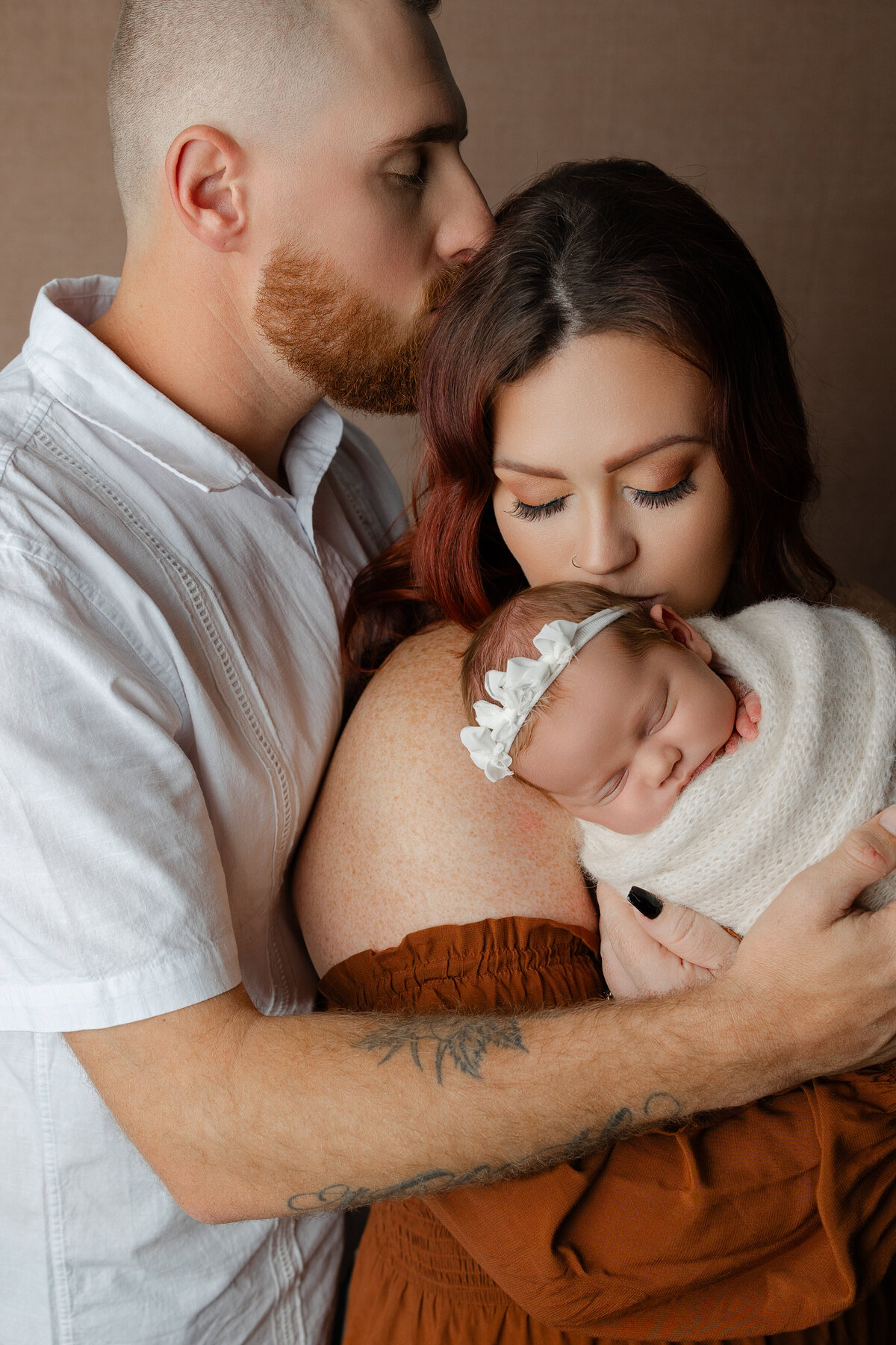 Mom and dad embracing their swaddled newborn baby girl, with dad kissing mom’s forehead and baby resting peacefully against mom’s chest. Photograph by Inland Empire newborn photographer Kaitlyn Dawn Photography.