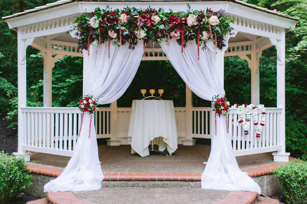 Ceremony gazebo with sheer white fabric and burgundy flowers.
