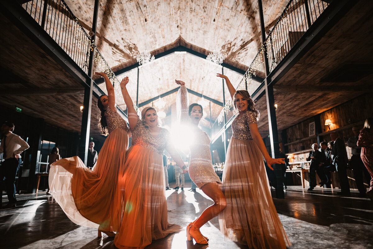 Bride and bridesmaids dancing under chandeliers at Hidden River Barns in the Lake District, wearing sparkling dresses in a stylish and rustic barn wedding venue.