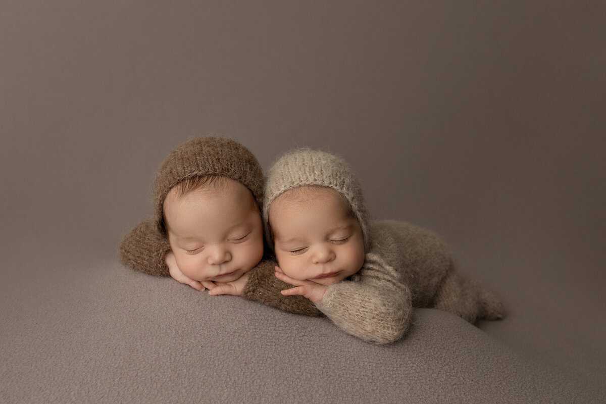Twin newborn photos by top London, Ontario photographer Amy Perrin-Ogg. Baby boys are wearing coordinating knit onesies and bonnets both laying on their bellies with their hands under their chin. One baby is resting his head and hand on his brother's shoulder.