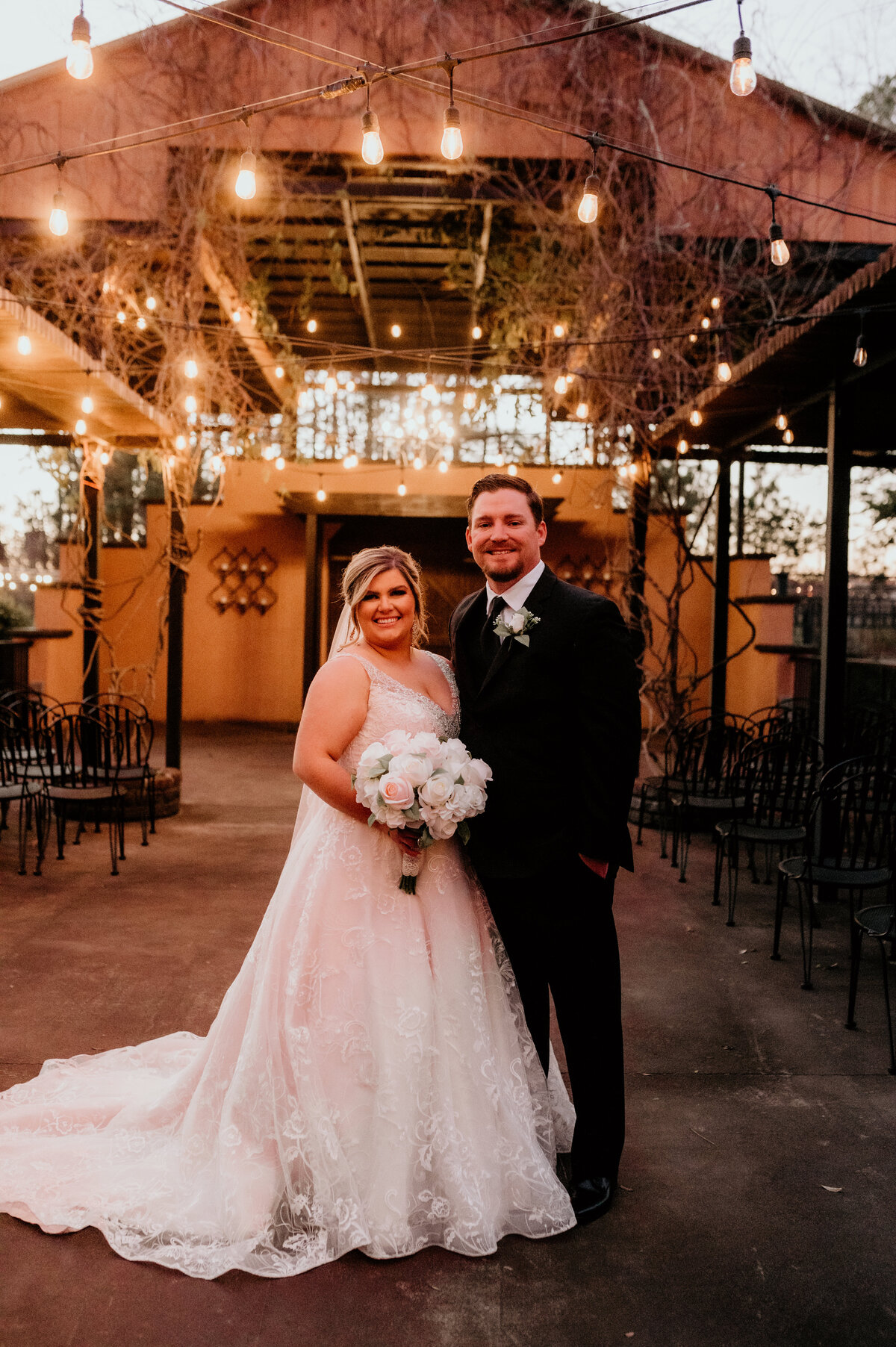 outdoor wedding reception space with string lights and stone walls with bride and groom standing together and smiling at the camera
