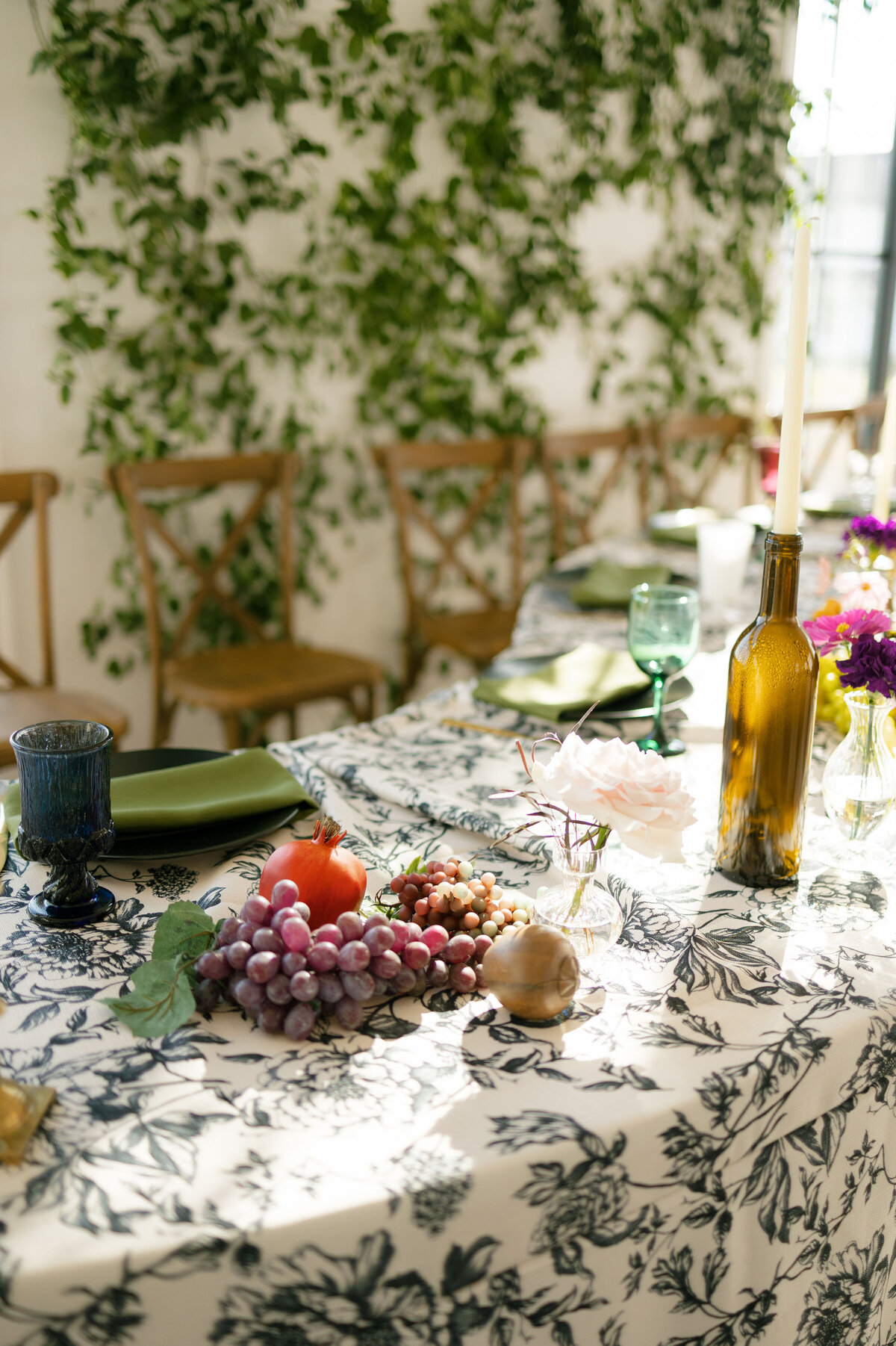 European-inspired wedding tablescape featuring black-and-white floral linens, wooden chairs, grapes, pears, figs, flowers in bud vases, and amber candlesticks, styled under natural sunlight for a romantic garden aesthetic.