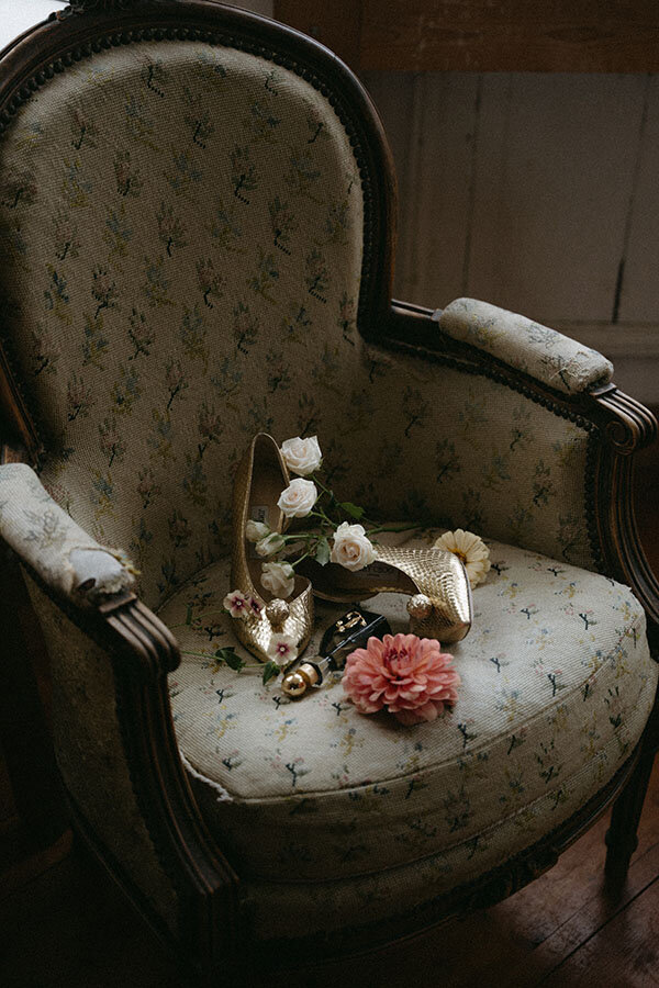Chaussures dorées, fleurs et parfum sur un fauteuil vintage. Détail capturé par Kamescop lors d'un mariage chic et rétro en Bretagne.