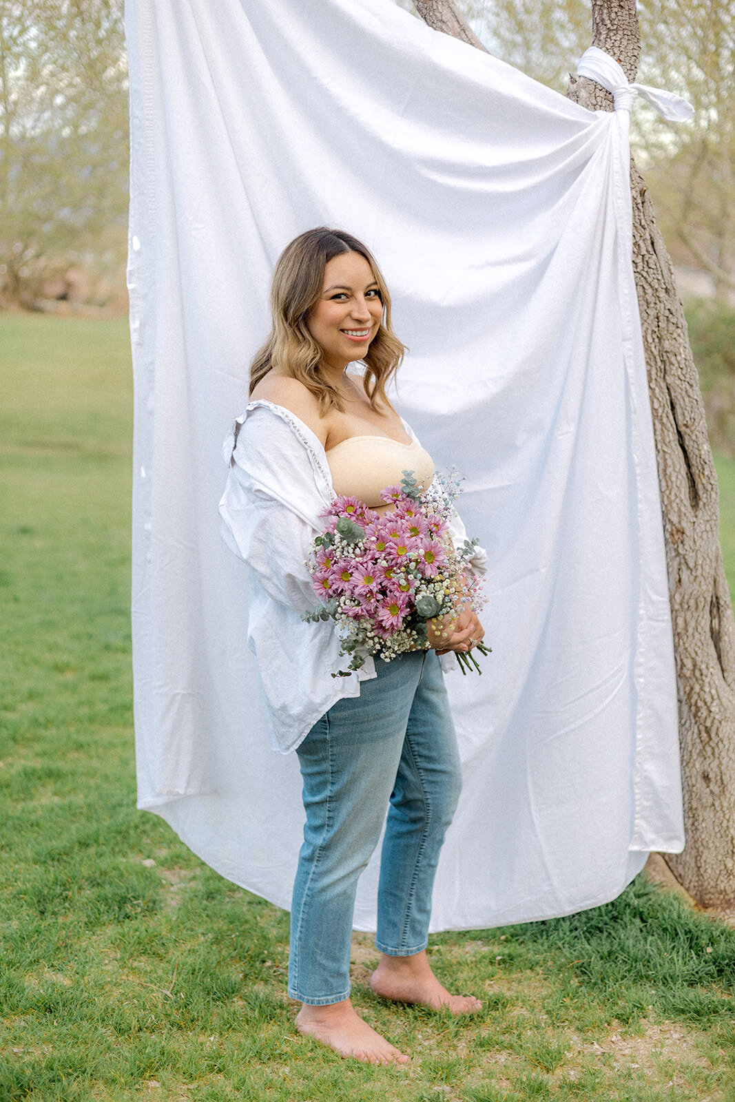 A pregnant woman stands in front of a sheet outdoors while holding flowers