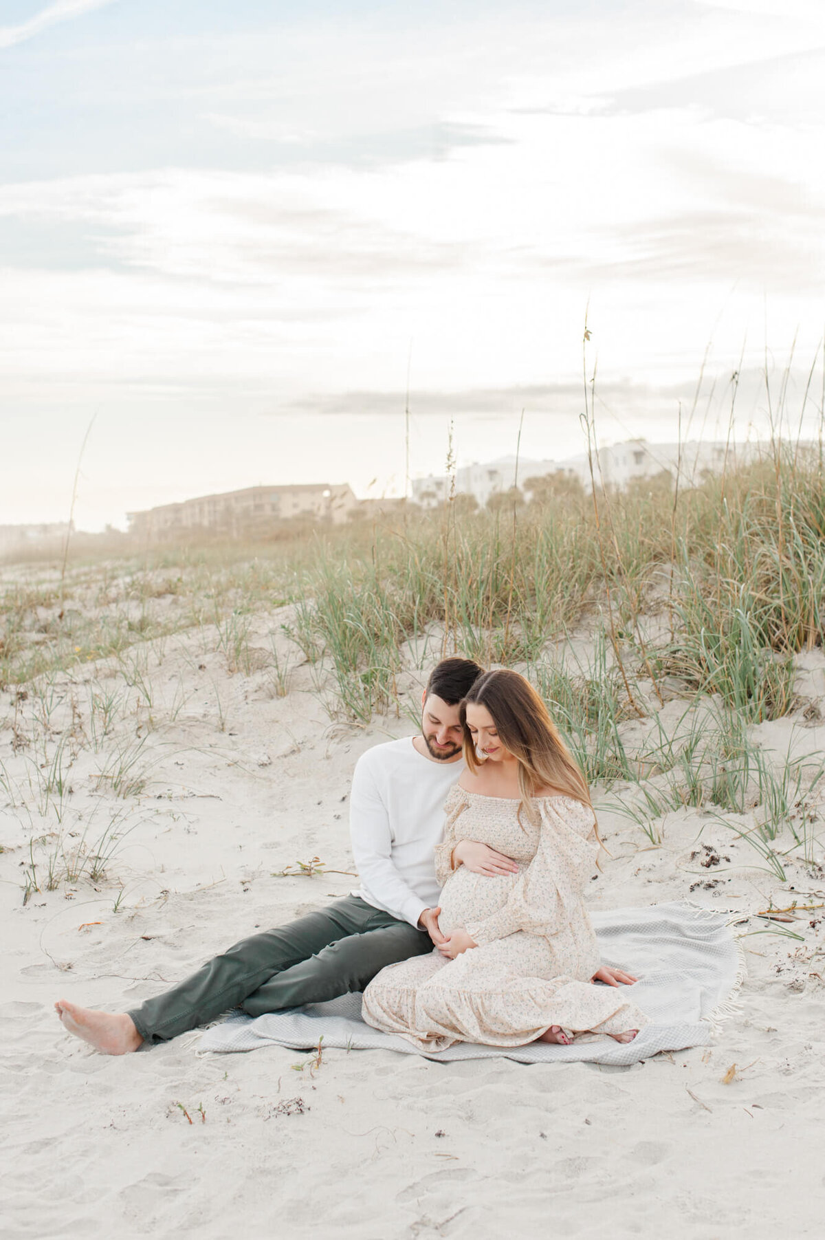 New parents sit in the dunes on a blanket and hold moms pregnant belly