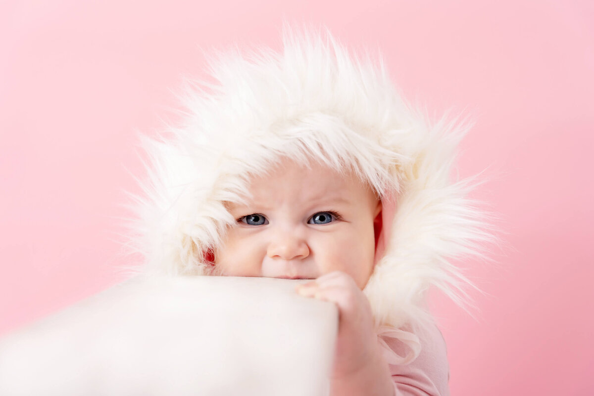 Baby girl wearing a white fuzzy hat bitting on a white ladder on a pink background for her 