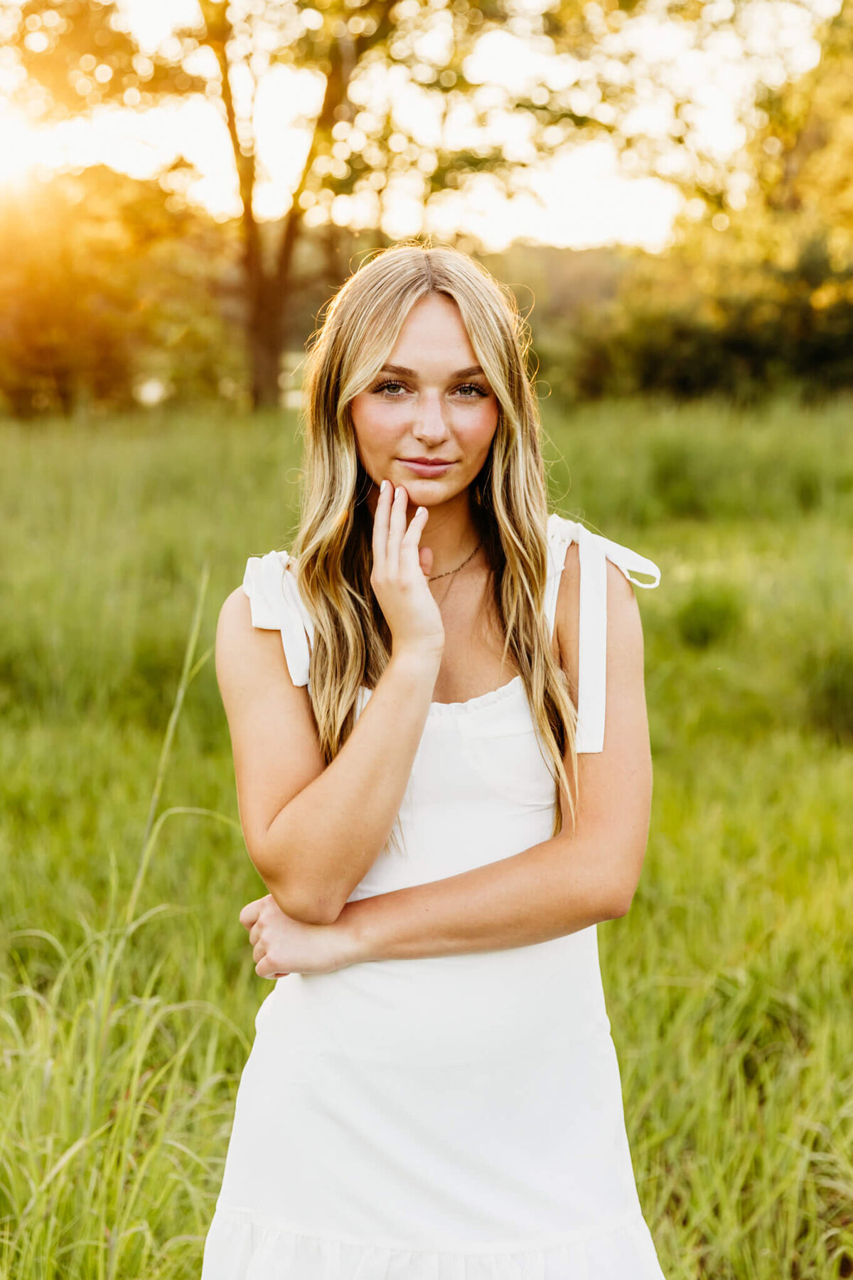 young woman looking serious in a white dress while standing in vibrant green grass for her high school senior photos by Ashley Kalbus