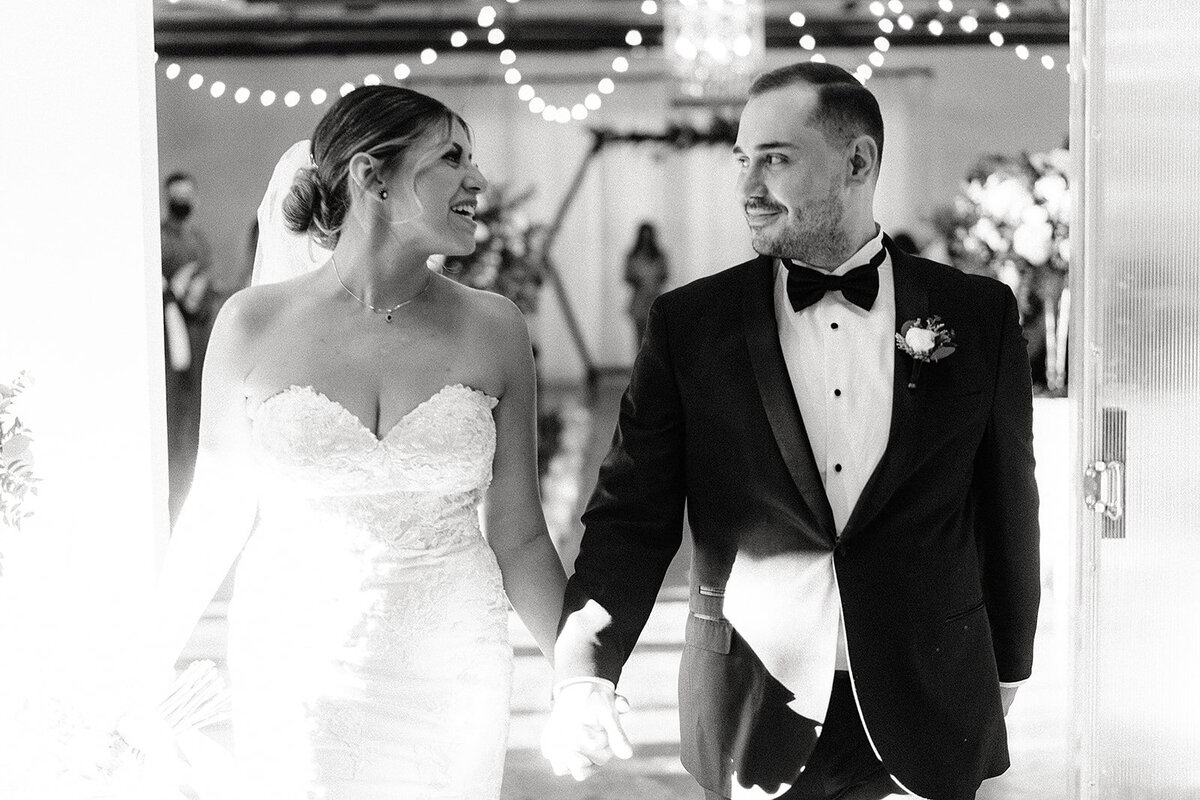 The newlyweds, smiling at each other, hold hands as they walk down the aisle lined with string lights and soft floral decorations.