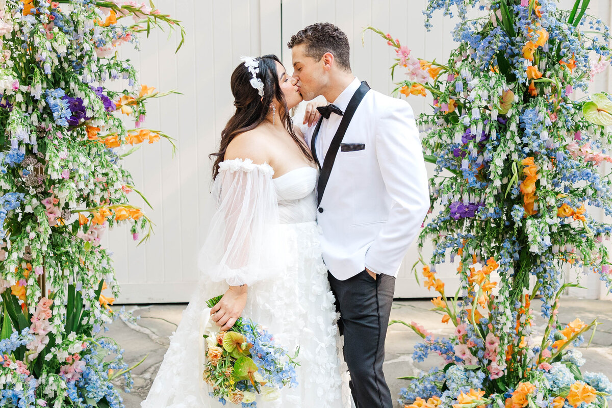 Celebrate love with this captivating photo of a couple sharing a kiss at their ceremony, surrounded by a delicate arch of wispy pastel and blue florals