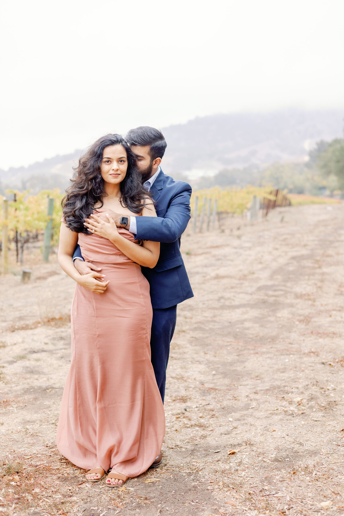 outdoor engagement portraits of man standing behind woman and leaning into kiss her neck as she smiles and holds his hands captured by bay area photographers